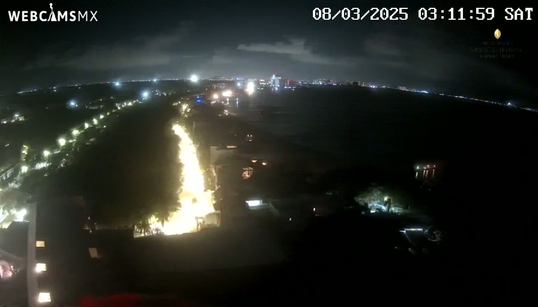 A nighttime, high-angle view shows a brightly lit coastal road winding through dark vegetation toward a city skyline across a body of water under a partly cloudy sky.