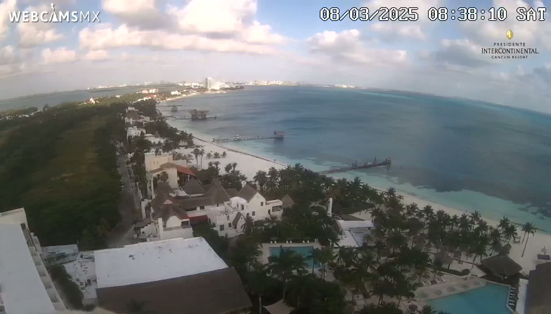 An aerial view shows a partly cloudy sky over a beach resort town with a white sand beach and calm, turquoise ocean.