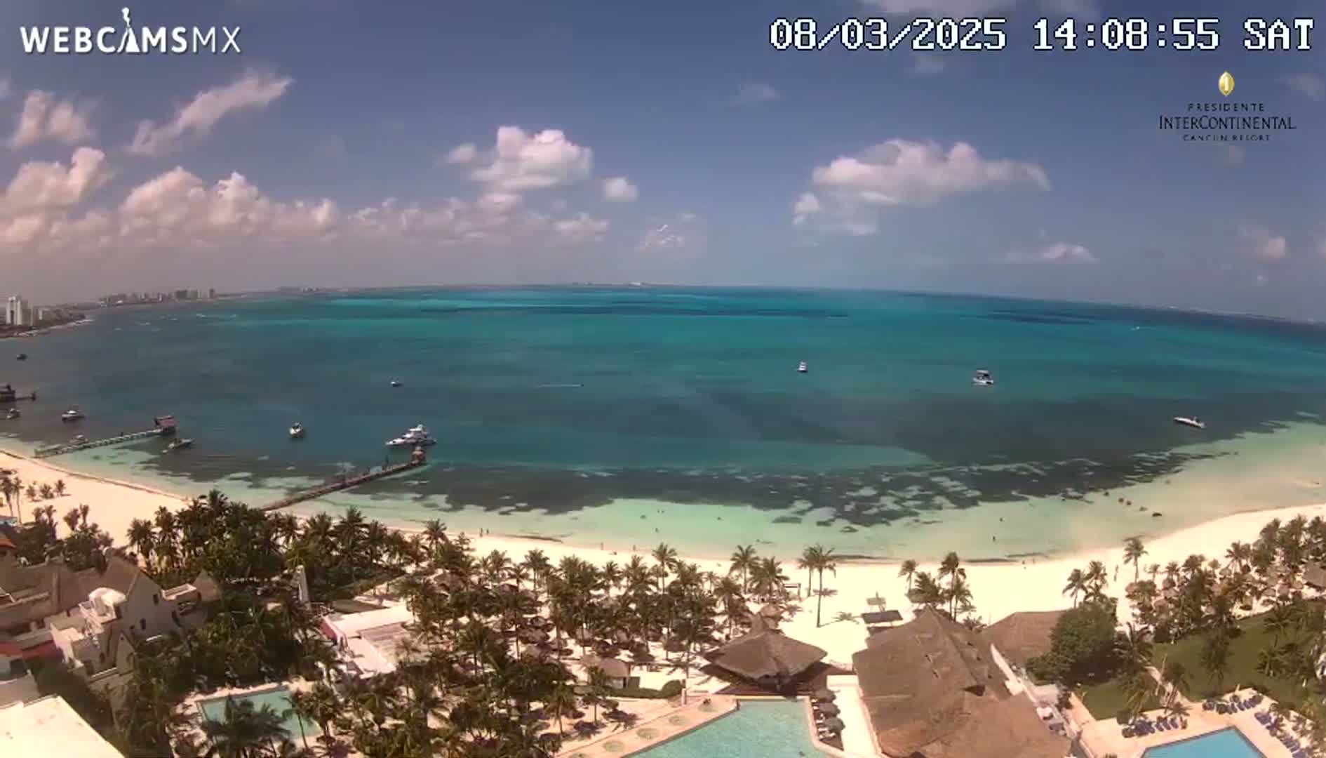 An aerial view shows a tropical beach with turquoise water, palm trees, a resort with swimming pools, and a few boats in the ocean under a partly cloudy sky.