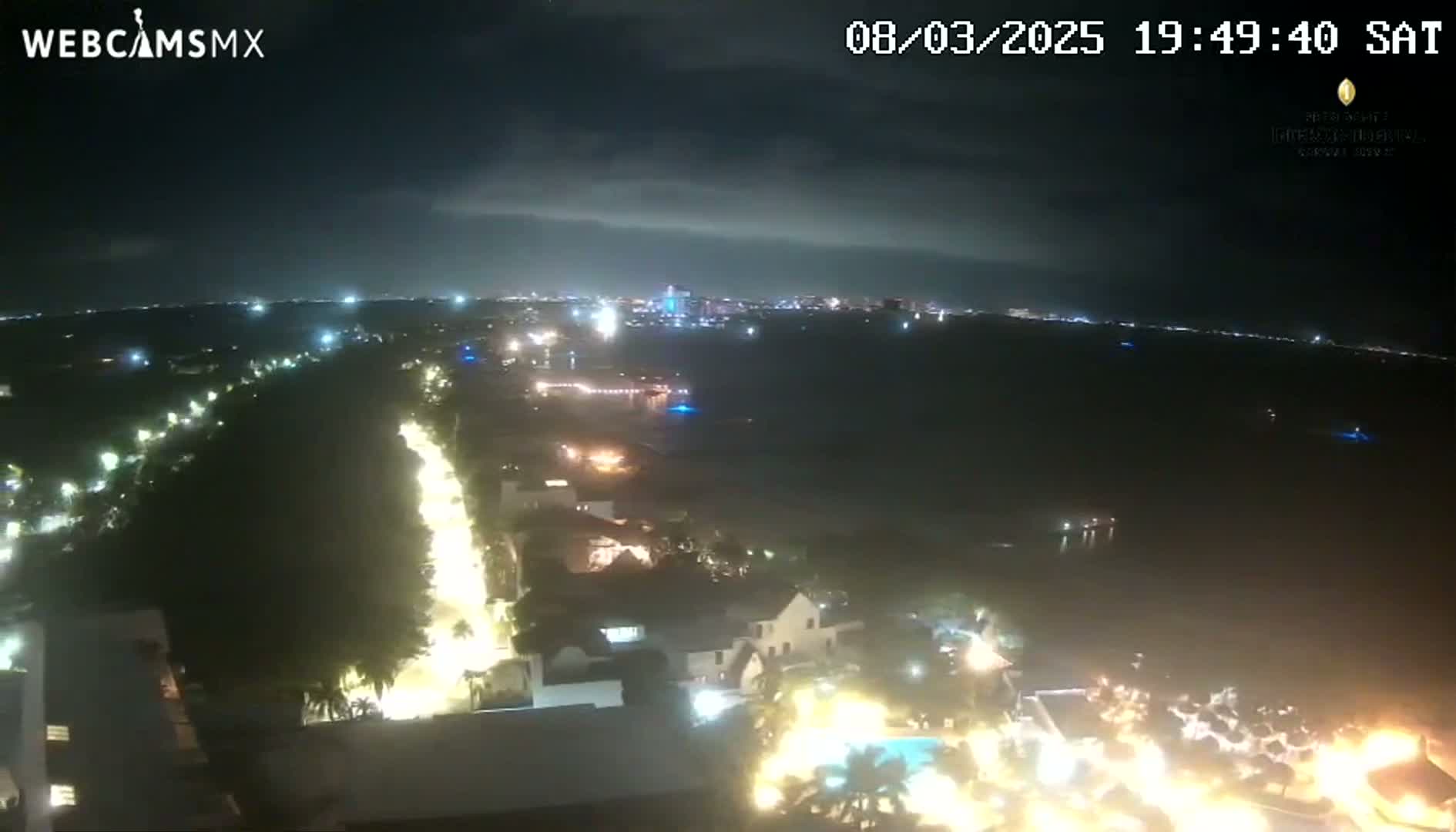 A nighttime, aerial view shows a coastal city with illuminated buildings and streets, under a partly cloudy sky.