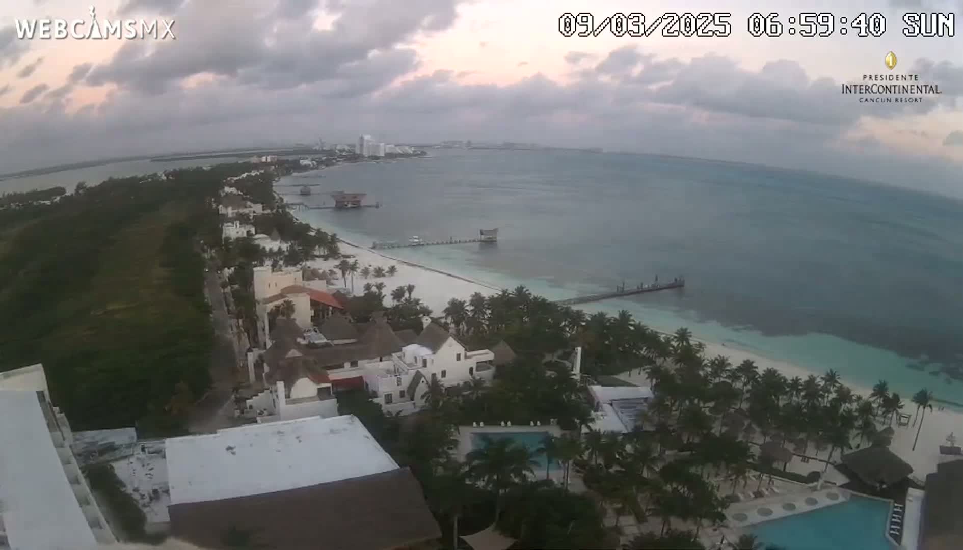 An aerial view of a beach resort on a partly cloudy morning, showing a long sandy beach, calm ocean, and tropical vegetation.