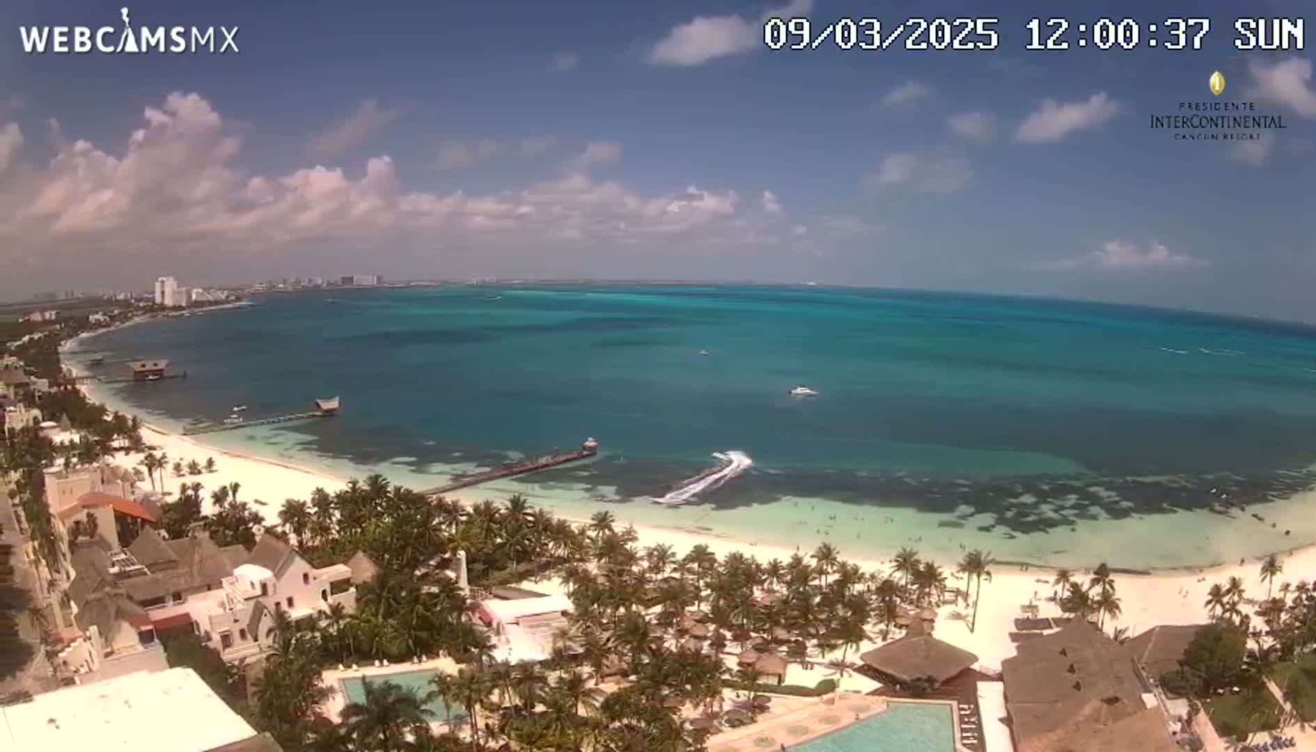 An aerial view shows a partly cloudy sunny day over a tropical beach with turquoise water, palm trees, and beachfront buildings.