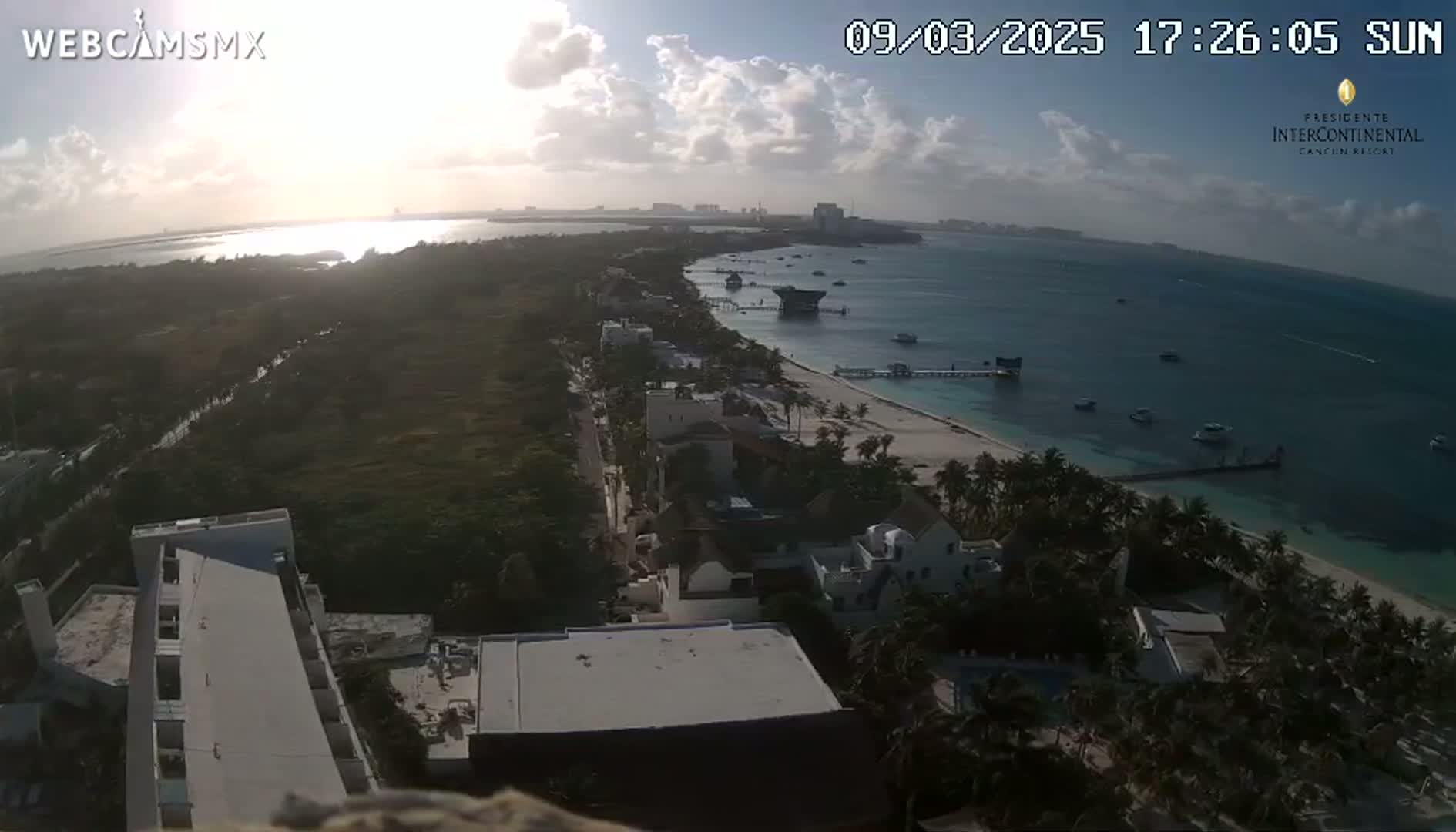 An aerial view shows a beach, ocean, and buildings under a partly cloudy sky at sunset.
