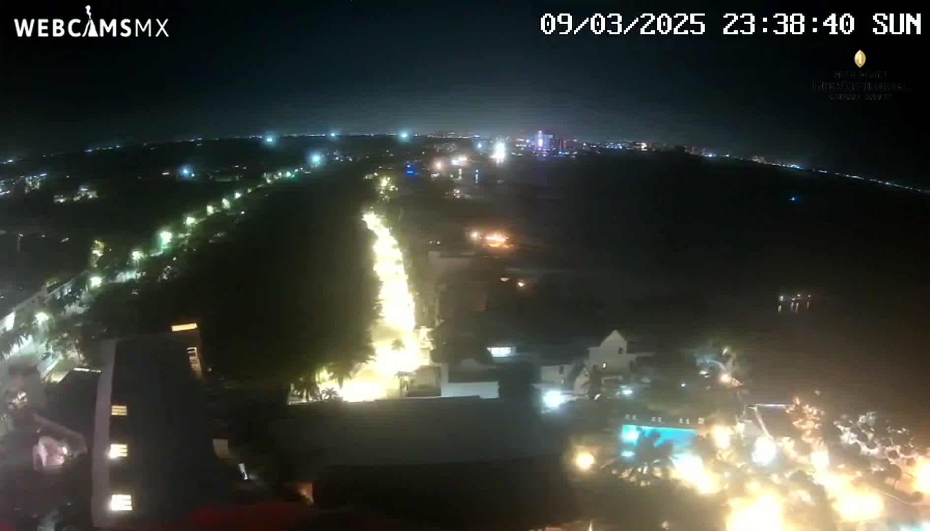A nighttime aerial view shows a brightly lit street cutting through a dark, tree-lined area, with buildings and pools of light visible nearby.