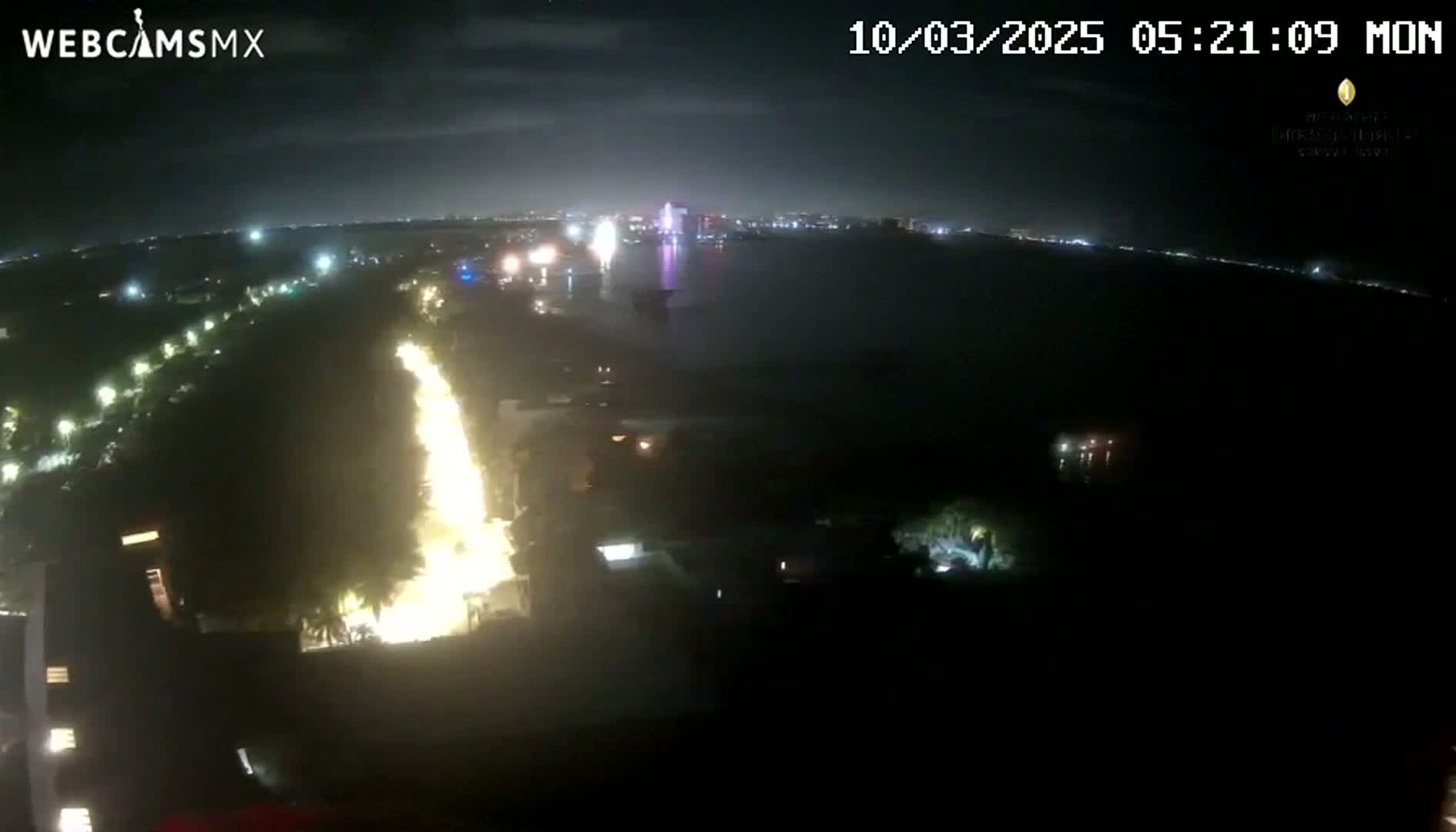 A nighttime aerial view shows a brightly lit roadway curving along a dark shoreline with fireworks visible in the distance over a body of water.