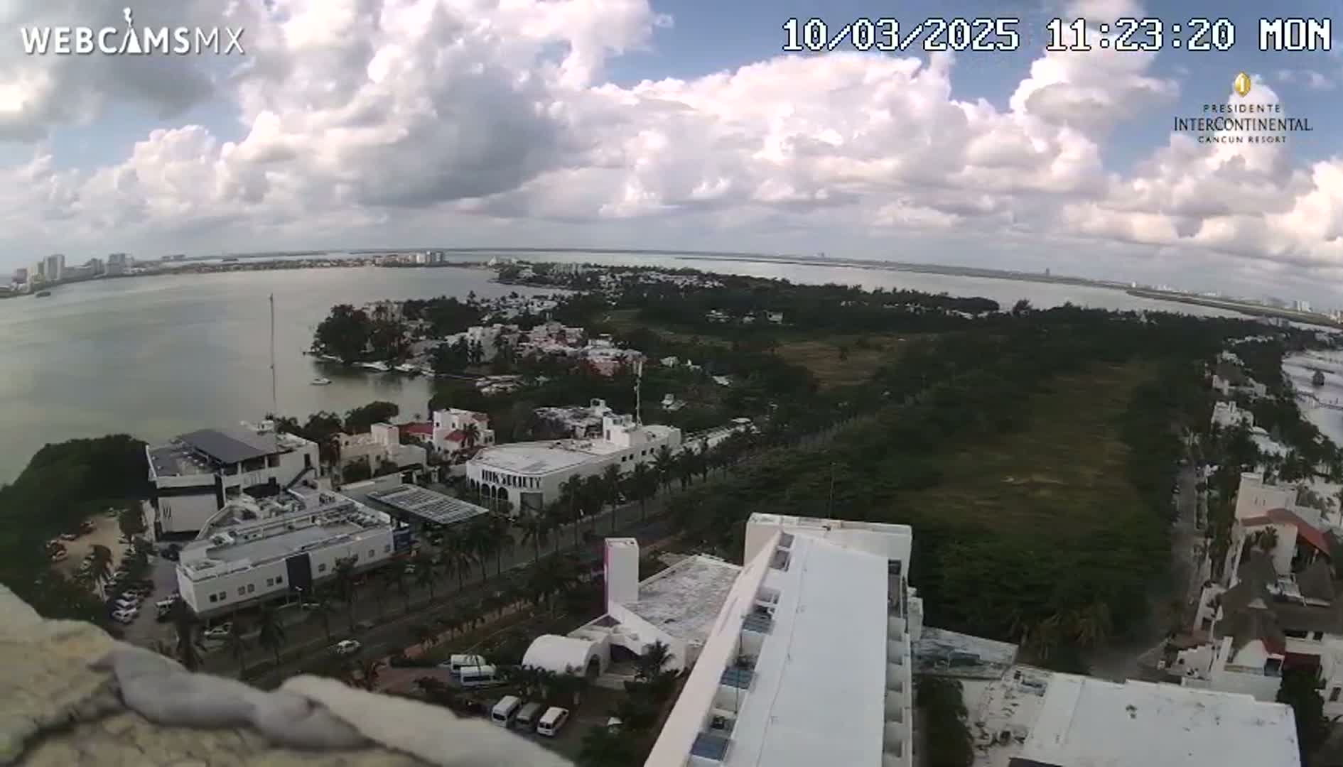 An elevated view shows a partly cloudy day over a coastal city with a body of water, buildings, and green areas.