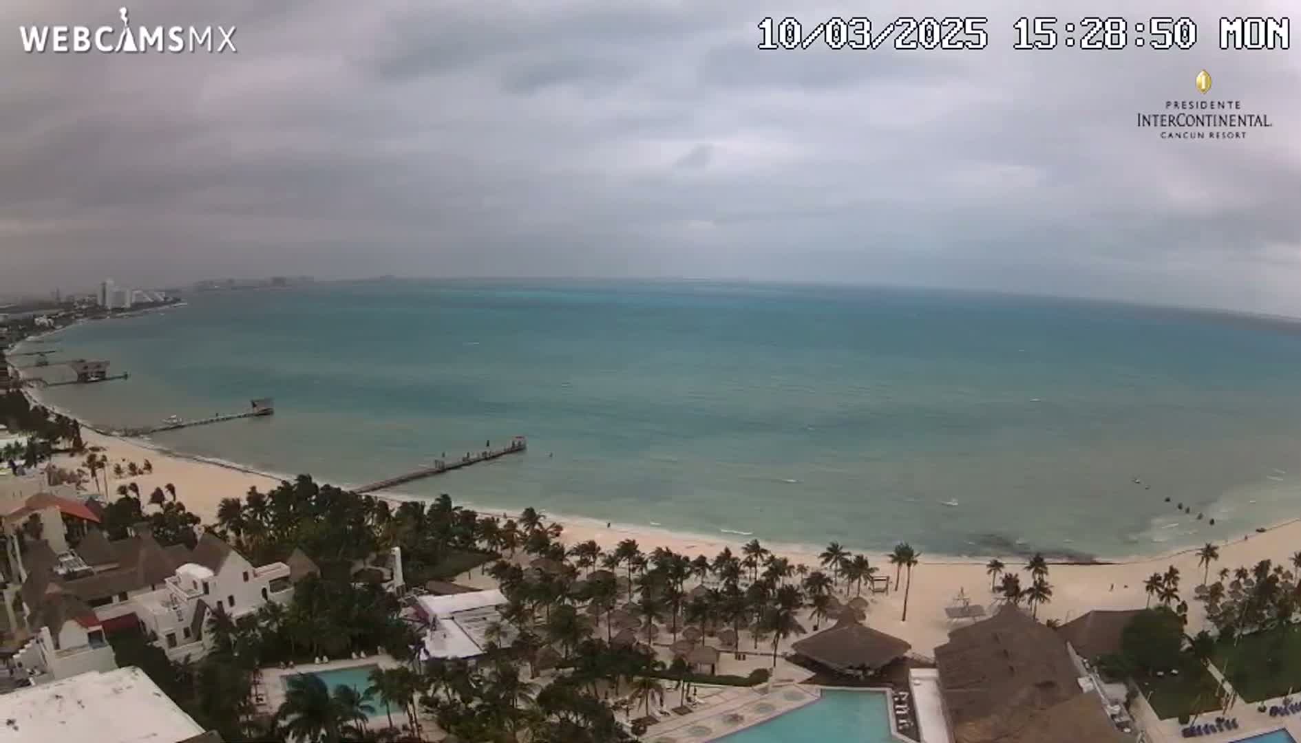 An aerial view shows a beach with calm, turquoise water under an overcast sky.