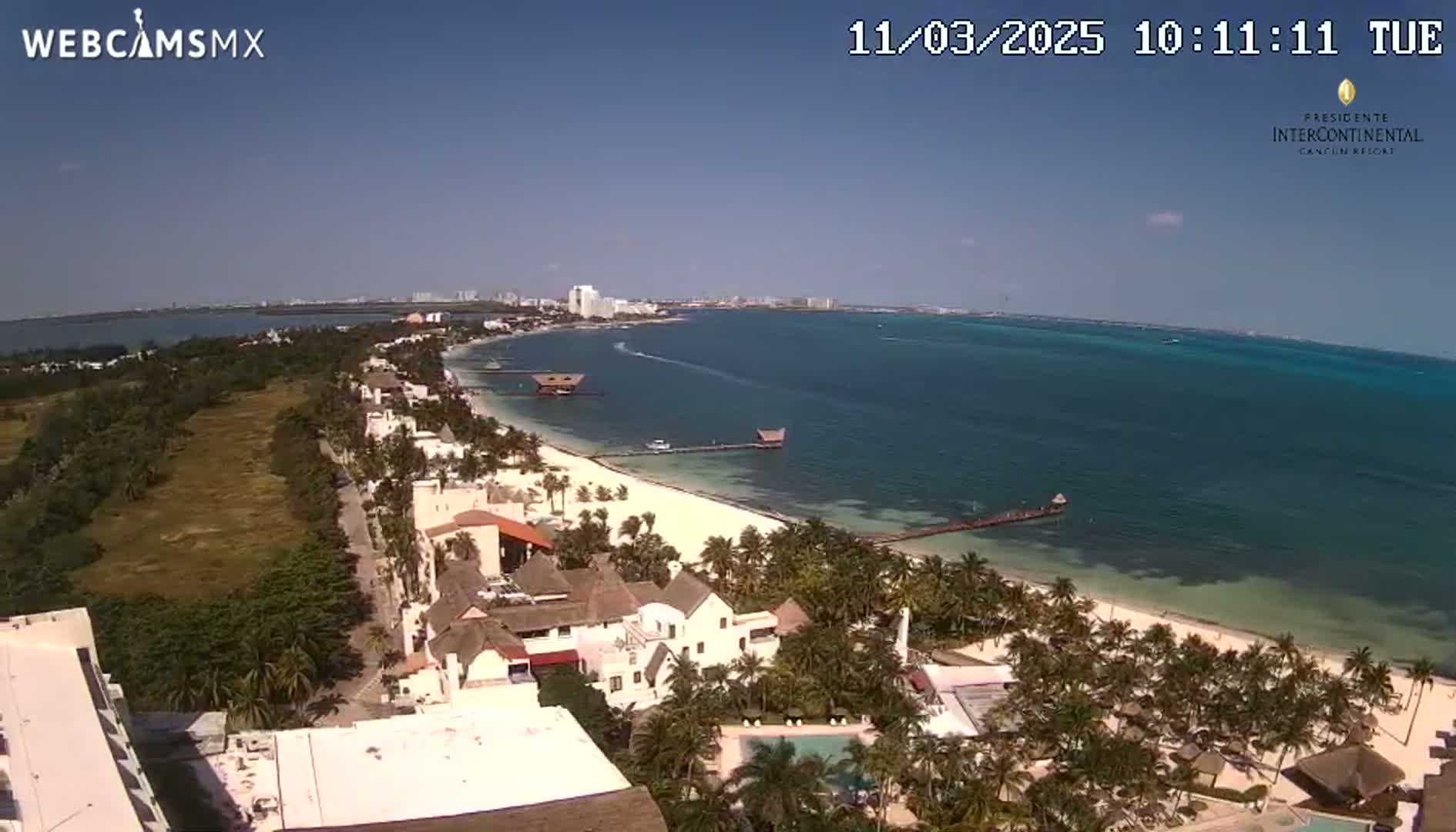 An aerial view shows a tropical beach with clear blue water, palm trees, and buildings under a sunny sky.