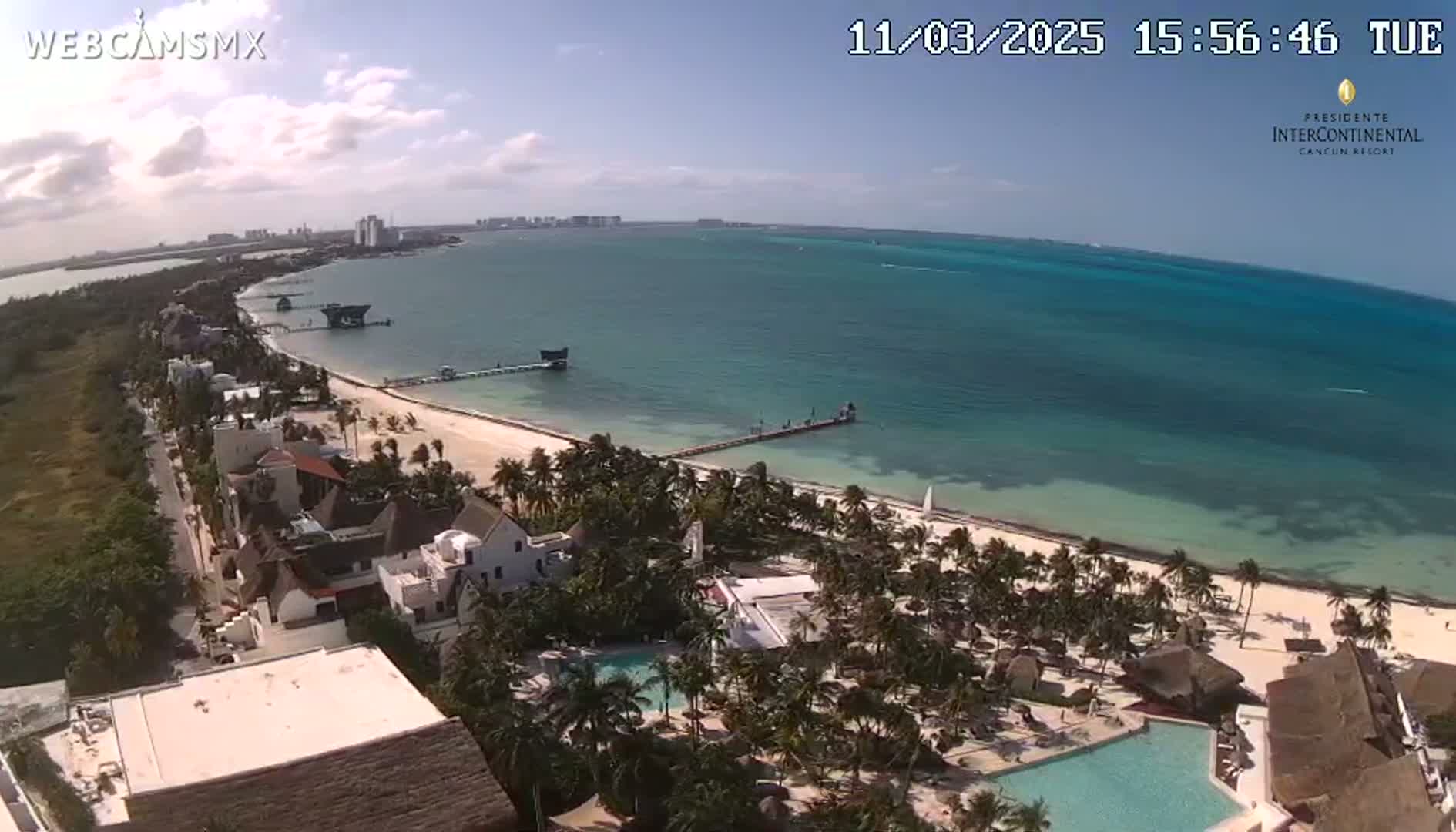 An aerial view shows a tropical resort with a beach and turquoise ocean under mostly sunny skies.