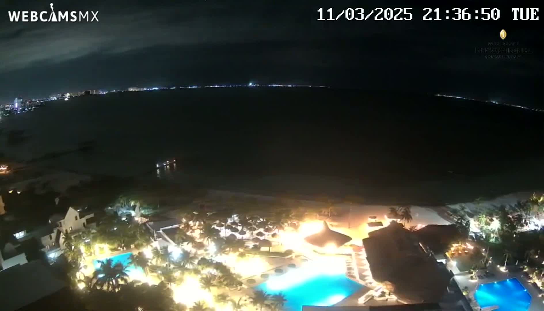 A nighttime aerial view of a resort complex with illuminated pools and buildings situated on a beach near a dark ocean under a mostly clear sky.