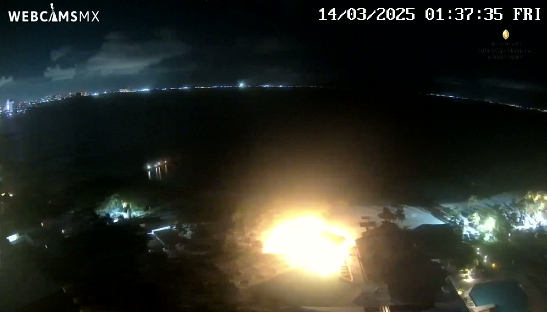 A large fire burns at night near a body of water with a city skyline visible in the distance under a partly cloudy sky.