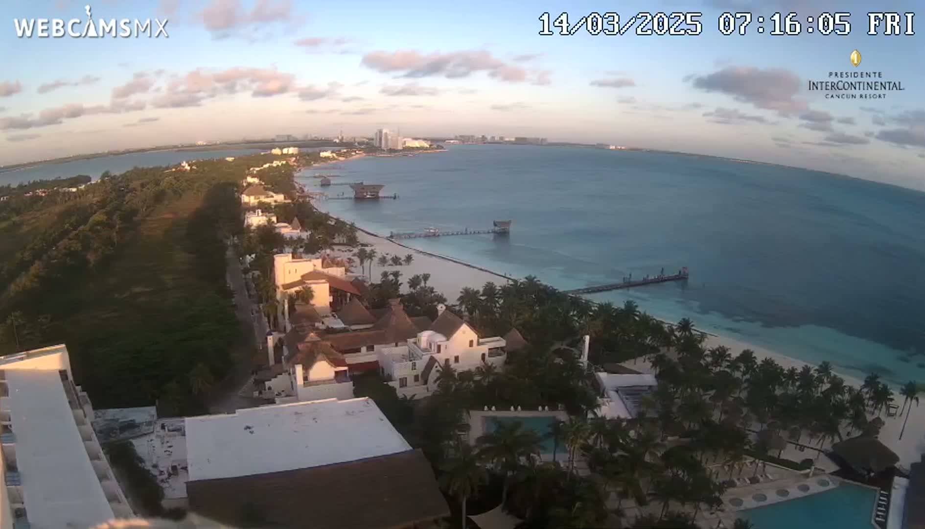 An aerial view shows a tropical coast at sunrise, with calm turquoise water, a sandy beach lined with palm trees, and resort buildings.