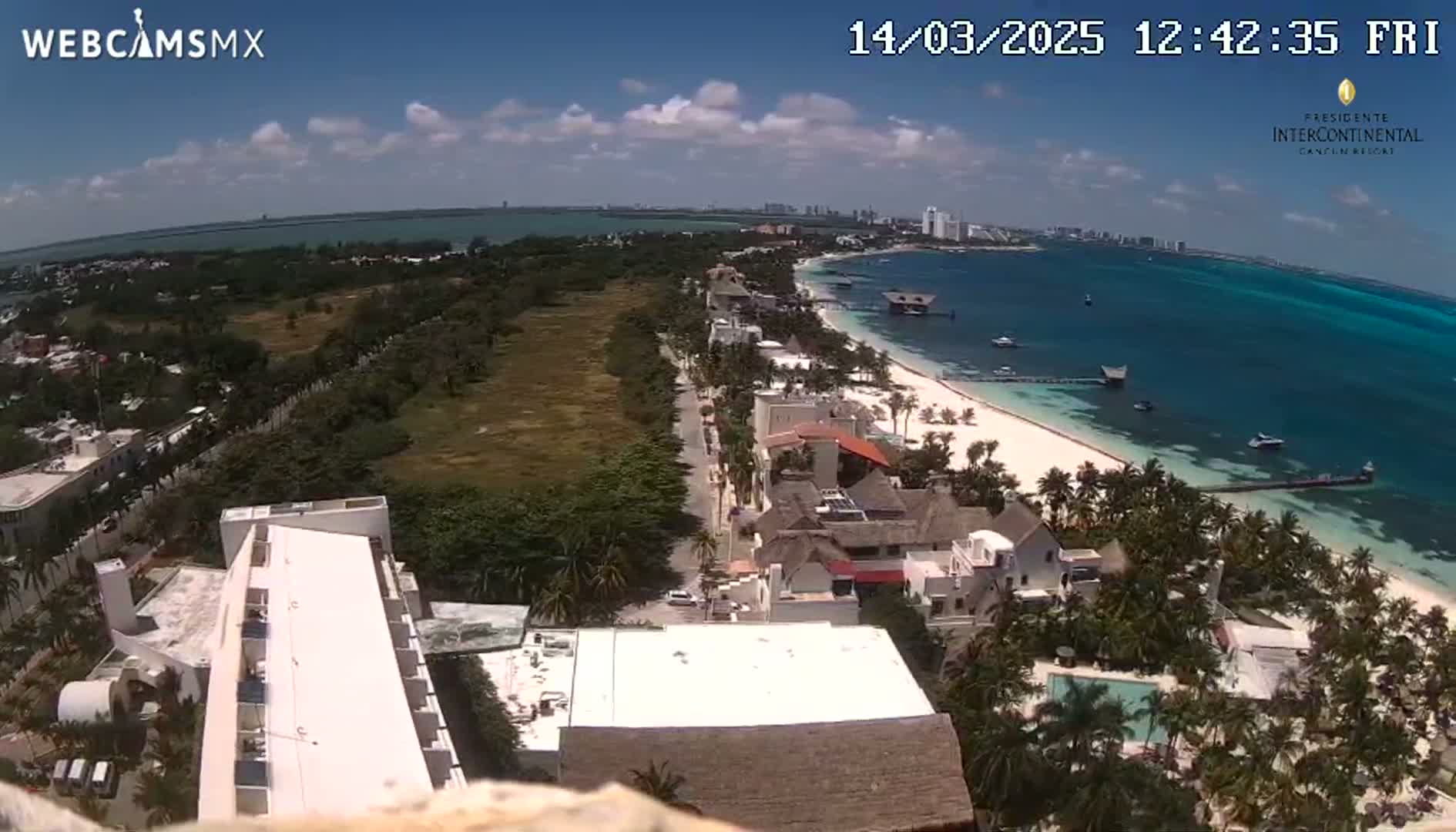 An aerial view shows a tropical beach with turquoise water under a partly sunny sky, featuring buildings, vegetation, and boats in the distance.