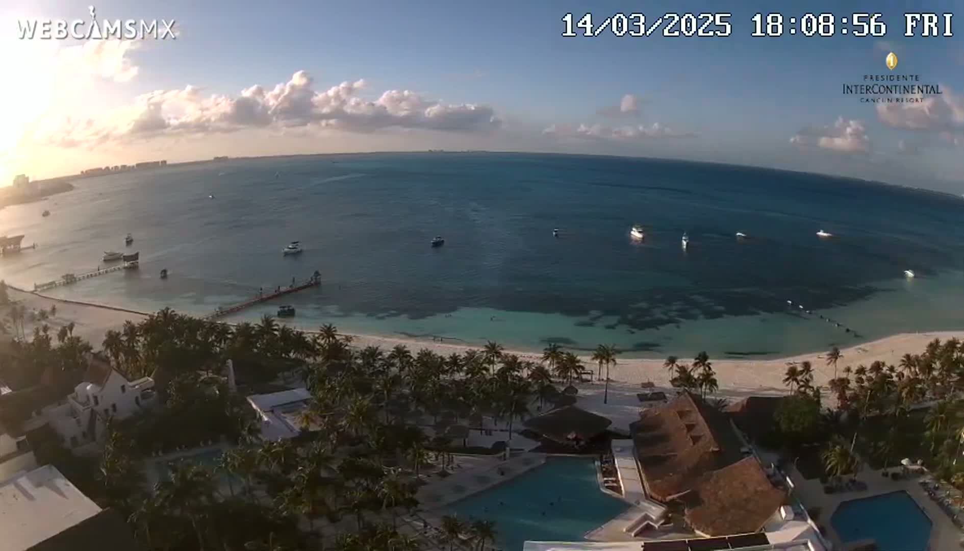 An aerial view shows a calm ocean with several boats, a white sandy beach lined with palm trees, and a resort with a swimming pool under a partly cloudy sky.