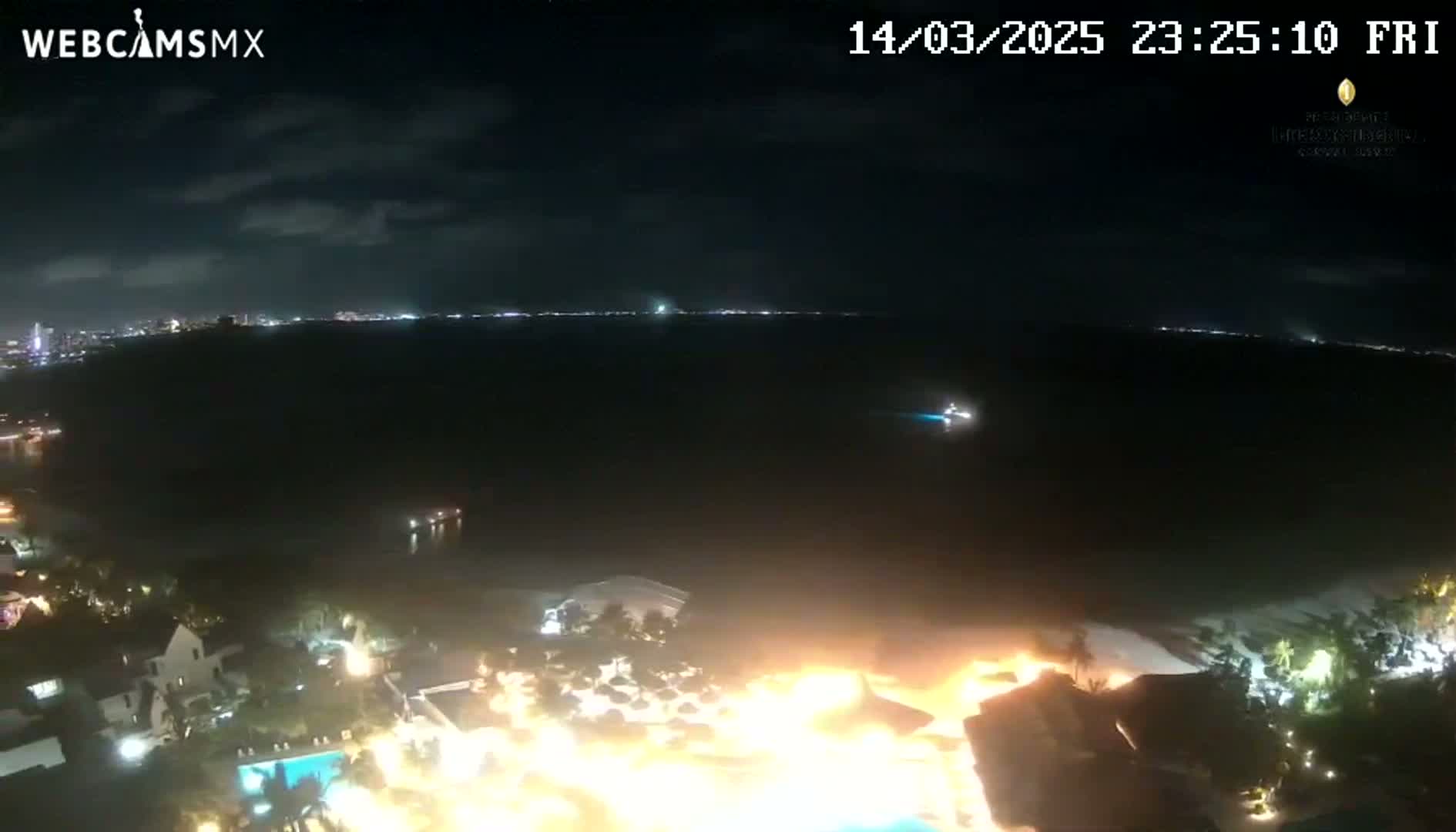 A large fire illuminates a resort area at night next to a dark ocean under a partly cloudy sky.