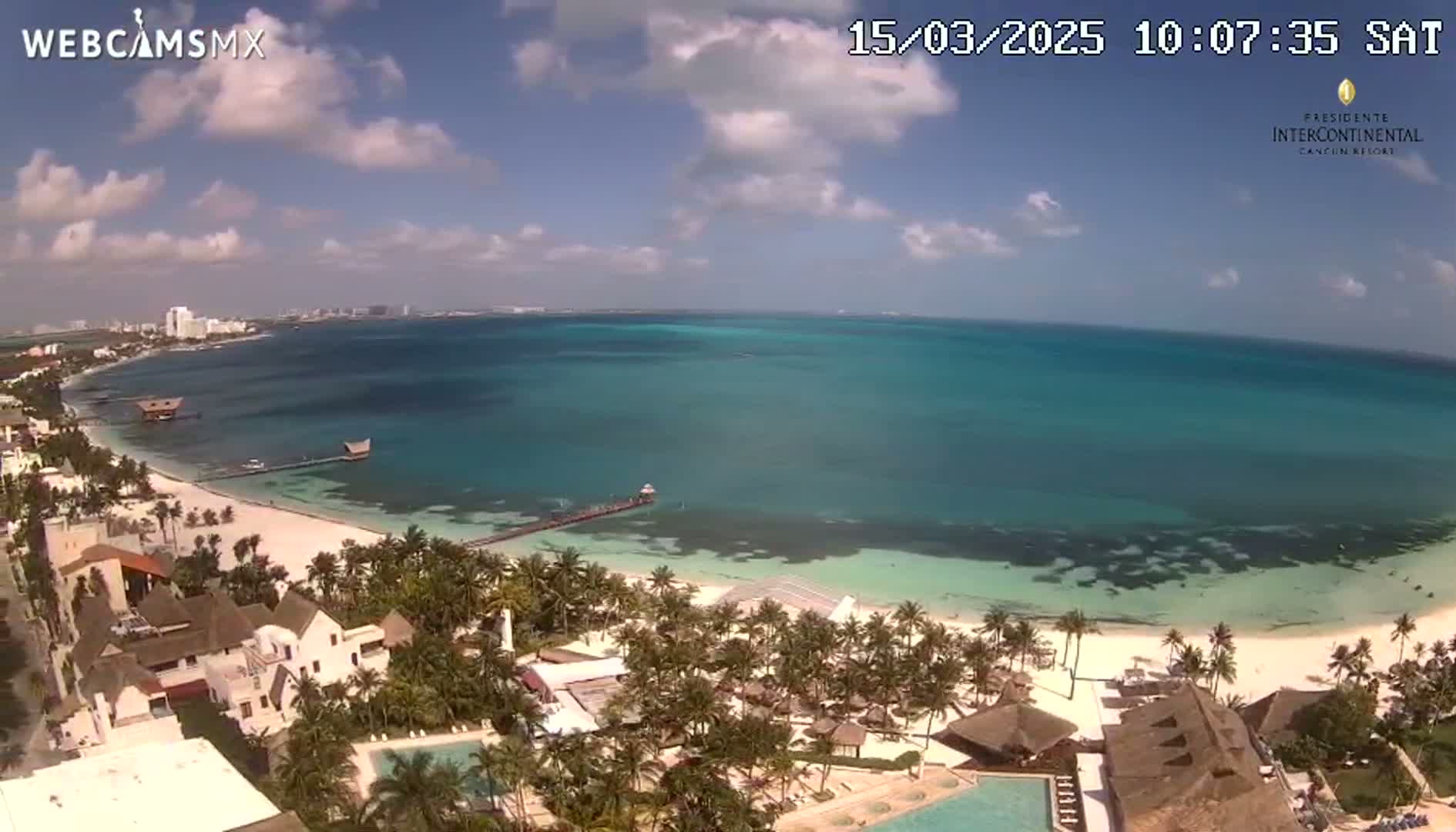 An aerial view shows a pristine beach with turquoise water under a partly cloudy sky.