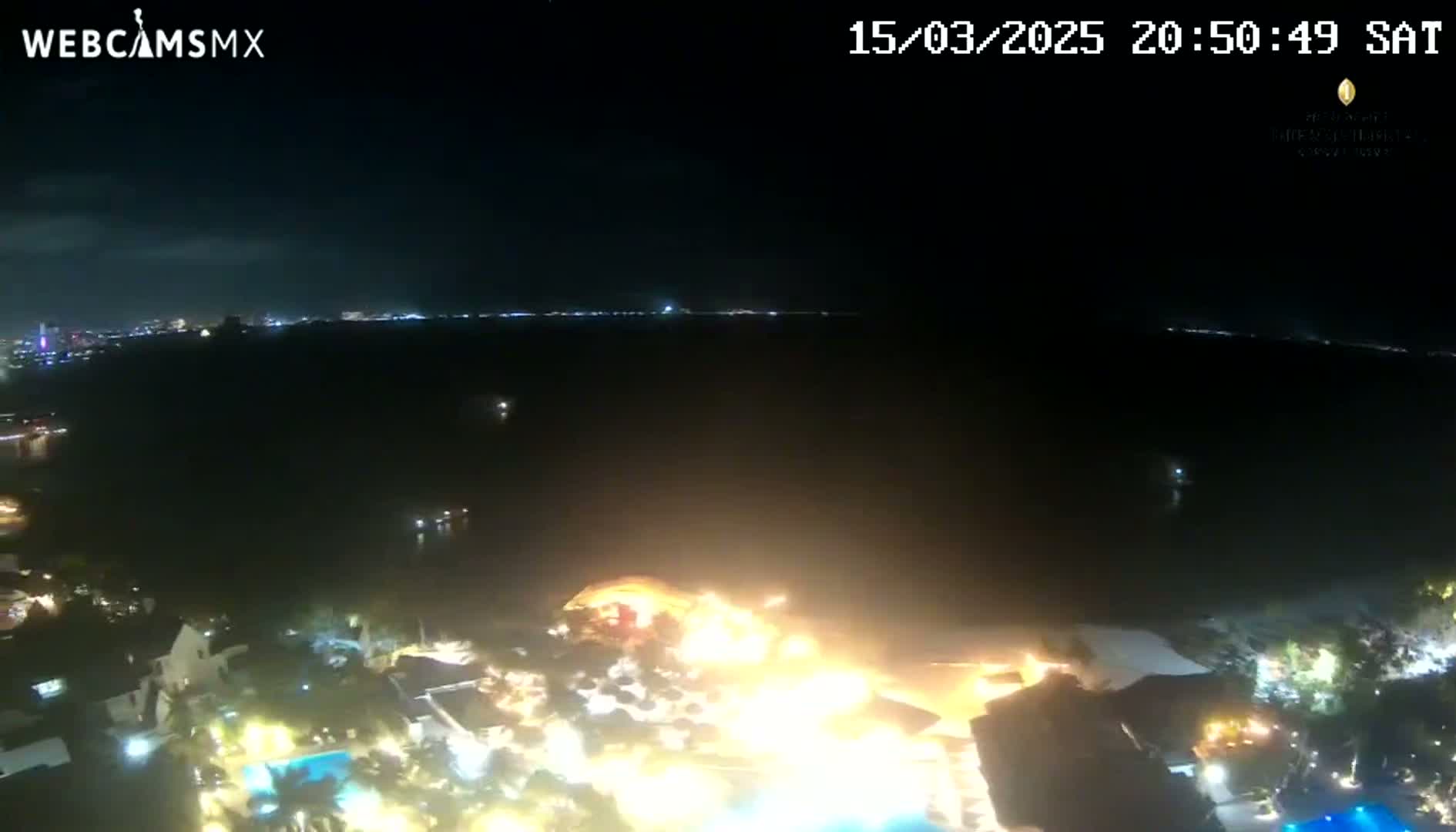 A nighttime, aerial view shows a dark body of water with a brightly lit city skyline in the distance and illuminated buildings and pools below.