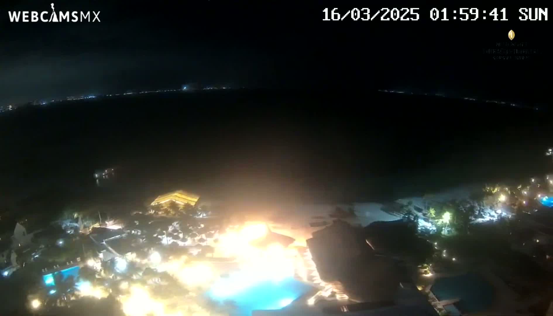 A nighttime aerial view shows a resort complex near a dark body of water, with a bright fire or intense light source near a pool.