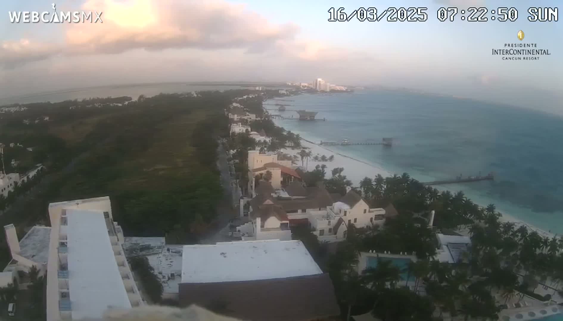 An aerial view of a beach resort town at sunrise, under partly cloudy skies, showing white sand beaches, turquoise water, and buildings nestled amongst palm trees.