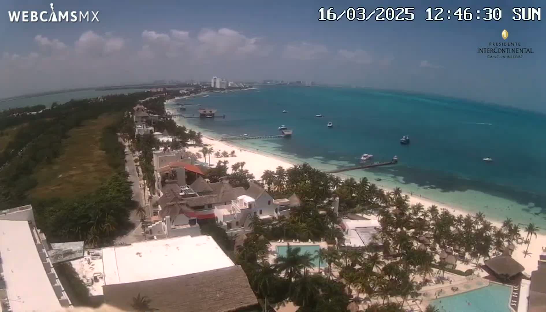A partly sunny day reveals a beachside resort town with a white sand beach, turquoise water, and palm trees under a mostly clear sky.