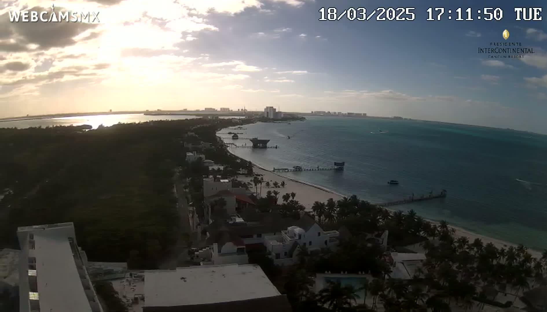 An elevated view of a beach at sunset, showing a calm ocean, a sandy shore lined with palm trees, buildings, and a pier extending into the water under partly cloudy skies.