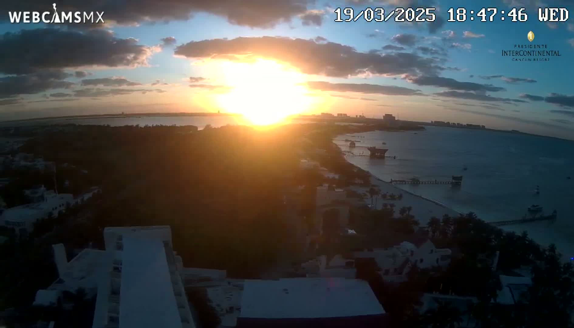 A coastal sunset over a calm body of water with a partially cloudy sky, viewed from a high vantage point overlooking buildings and a beach.
