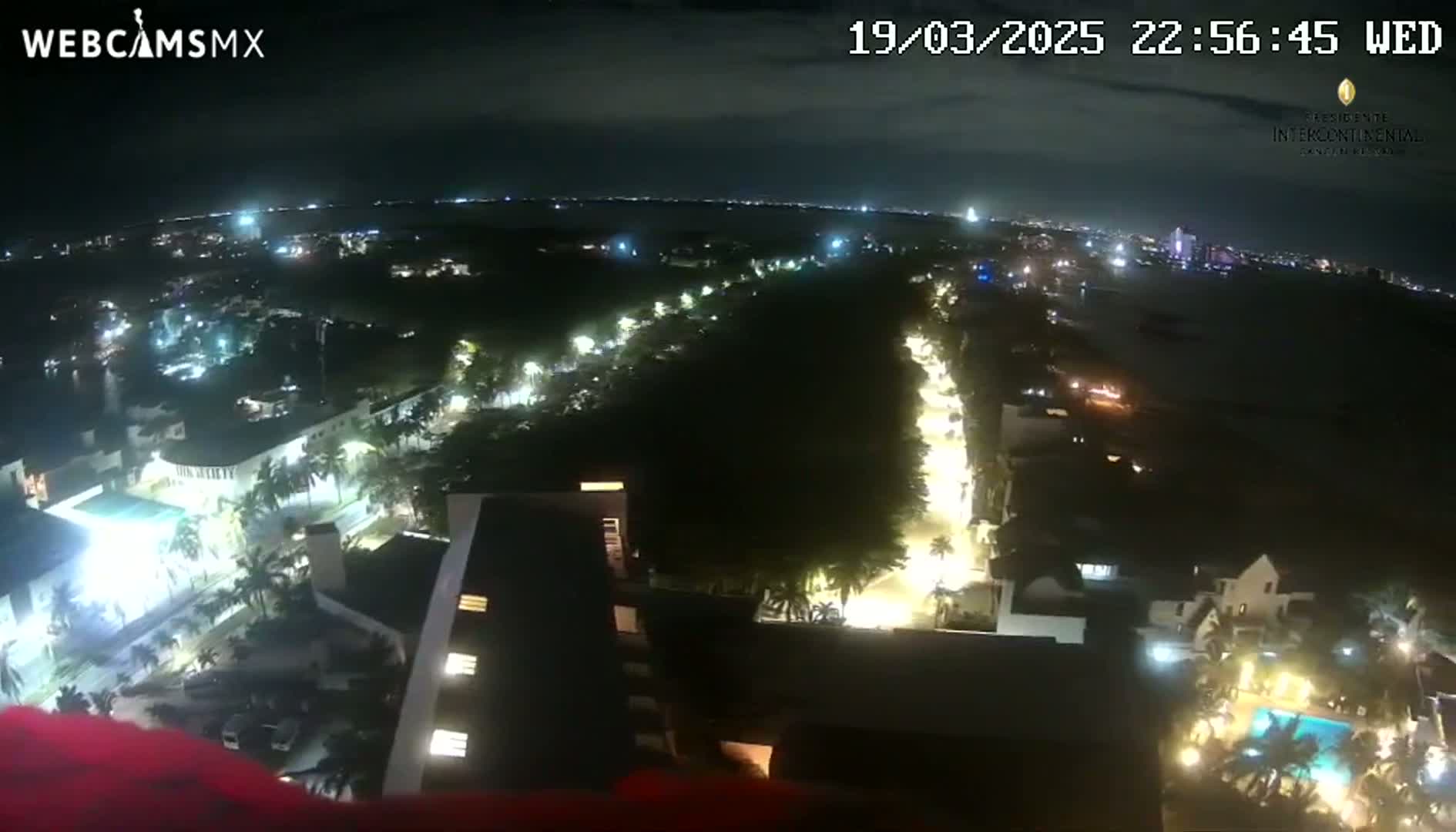 A nighttime, high-angle view shows a city illuminated by streetlights, with a body of water visible in the distance under a clear sky.