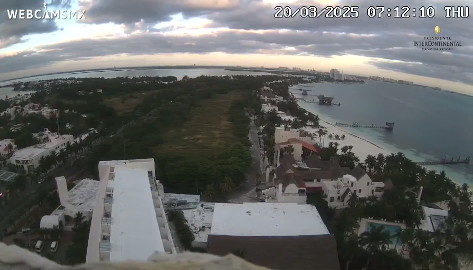 An elevated view shows a beach, ocean, and coastal buildings under a cloudy sky.