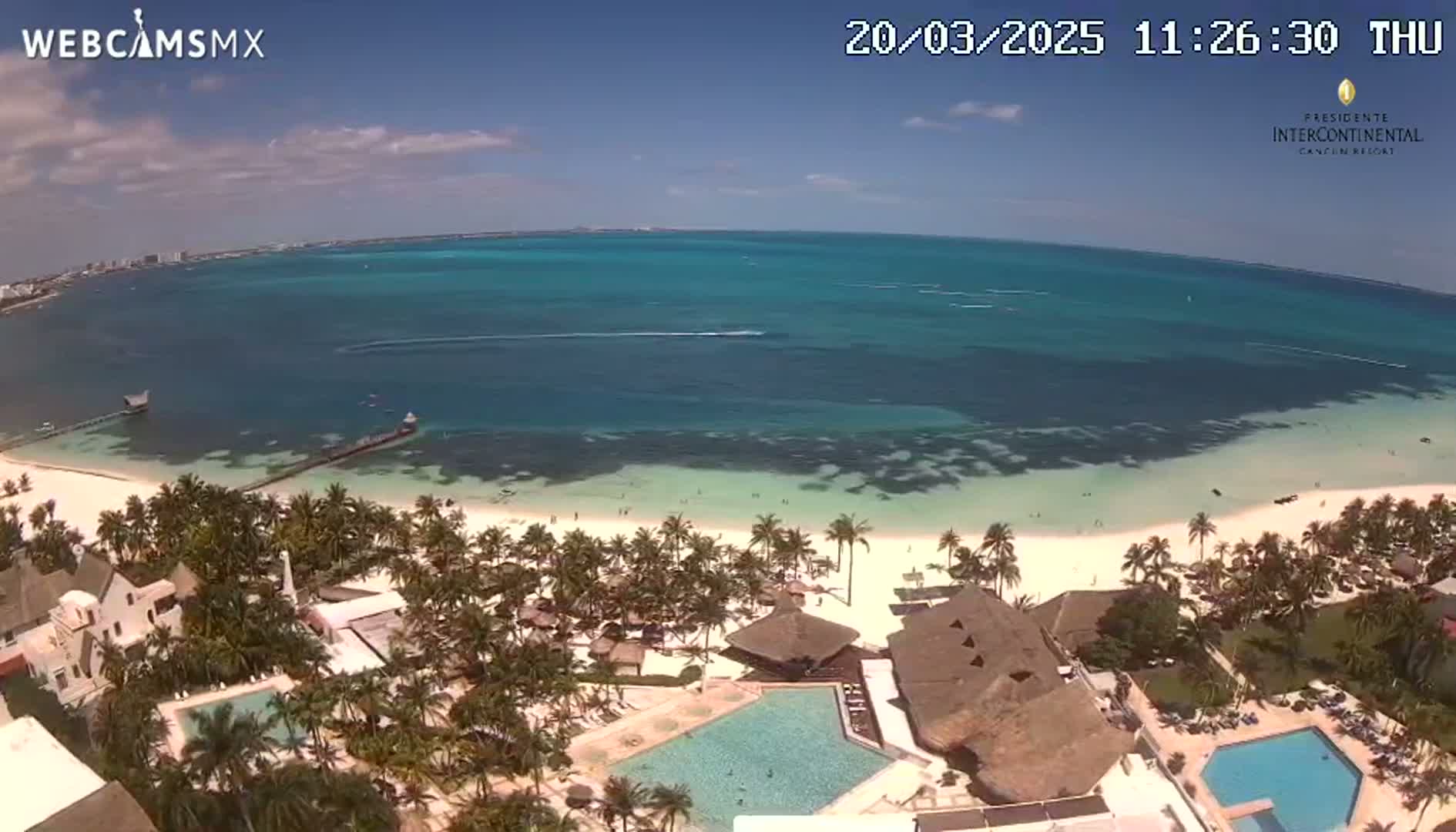 An aerial view shows a resort on a white sand beach with turquoise water under a mostly sunny sky.