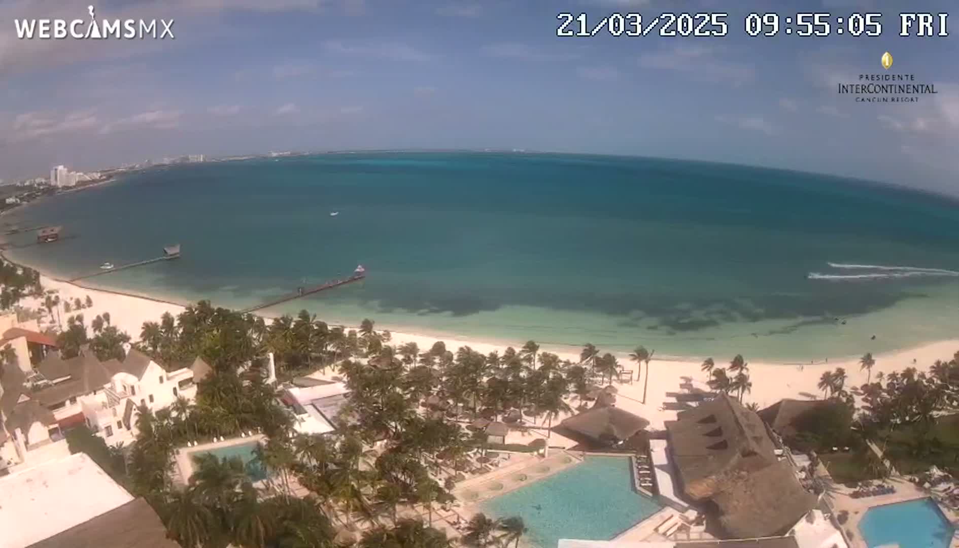 An aerial view shows a clear, sunny day at a tropical beach resort with calm, turquoise water, a white sandy beach lined with palm trees, and several resort buildings with swimming pools.