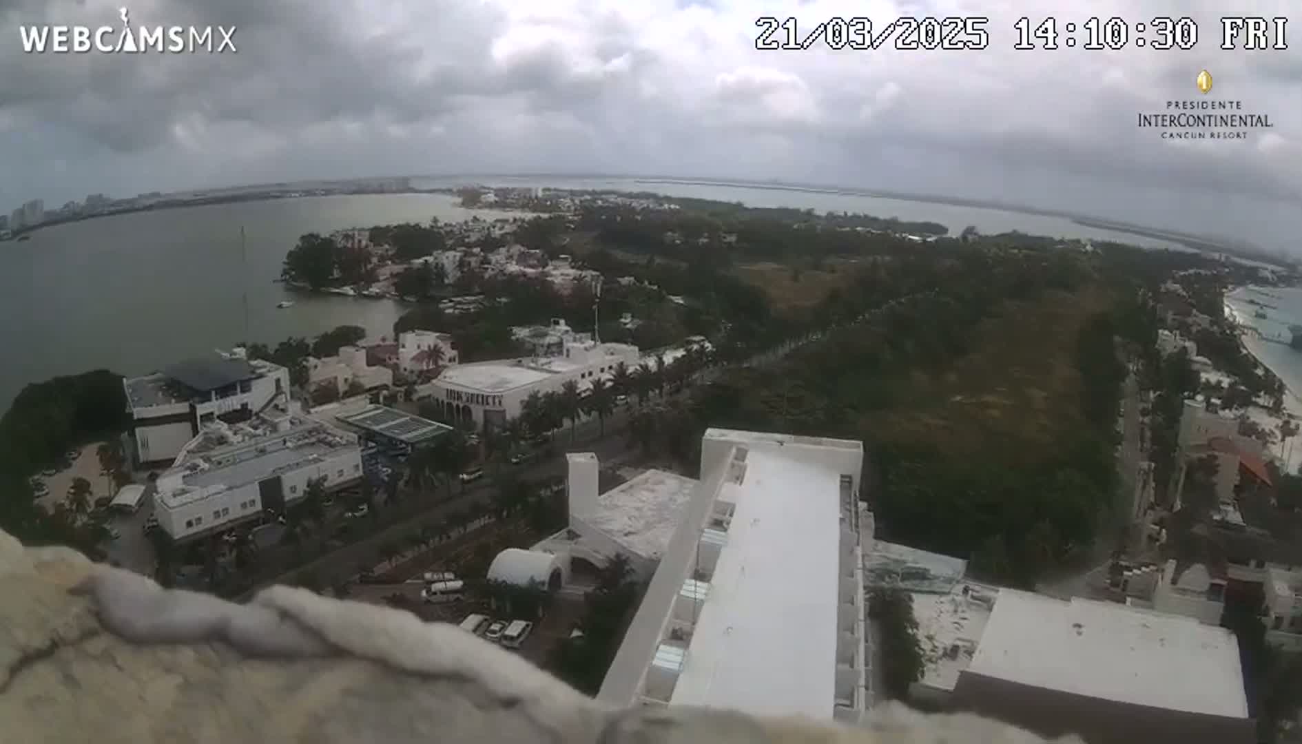 A high-angle view of a coastal city under a cloudy sky, showing buildings, a body of water, and lush green vegetation.
