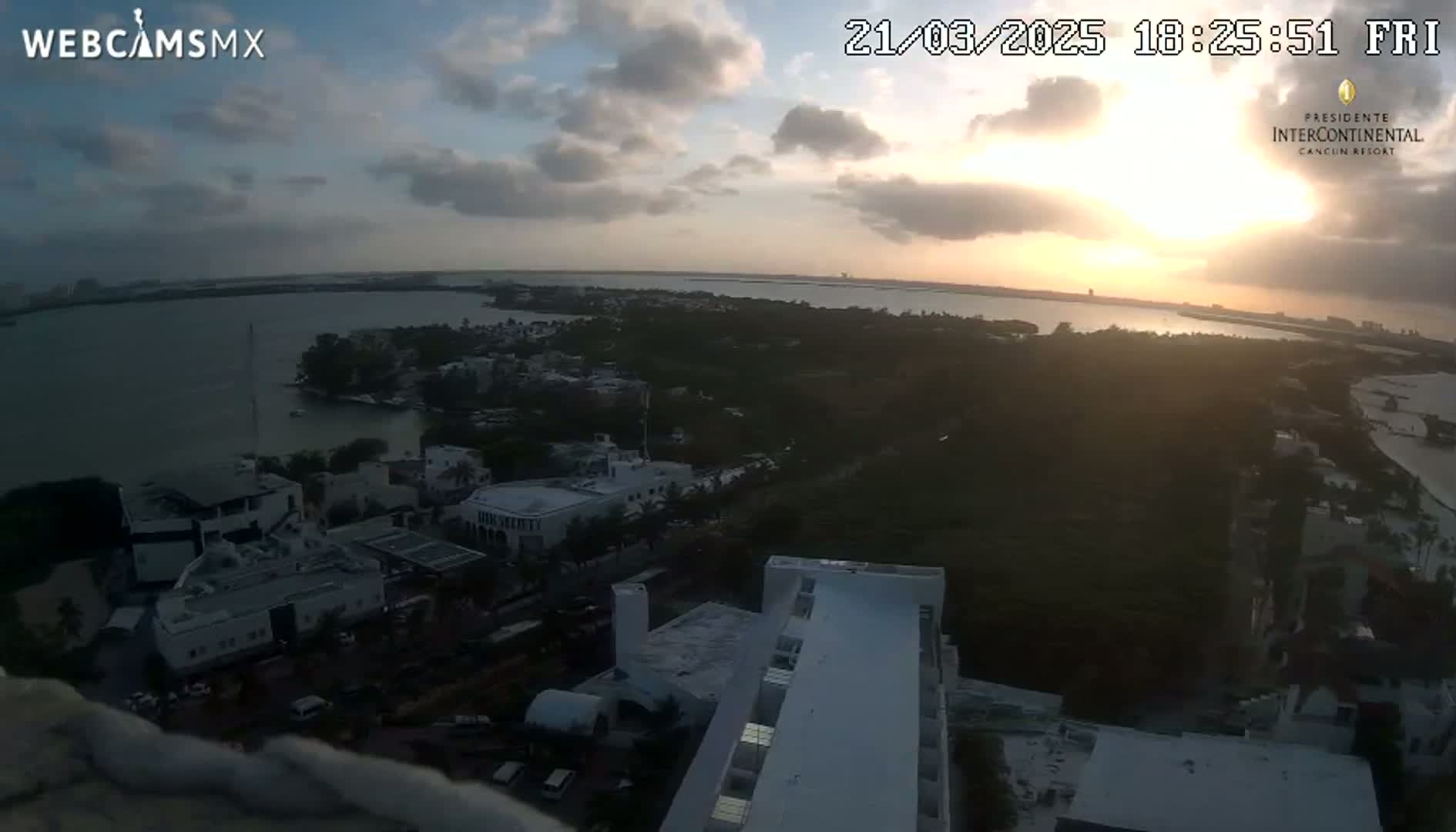 A sunset over a coastal town with a body of water, partially cloudy skies, and a view of buildings from a high vantage point.