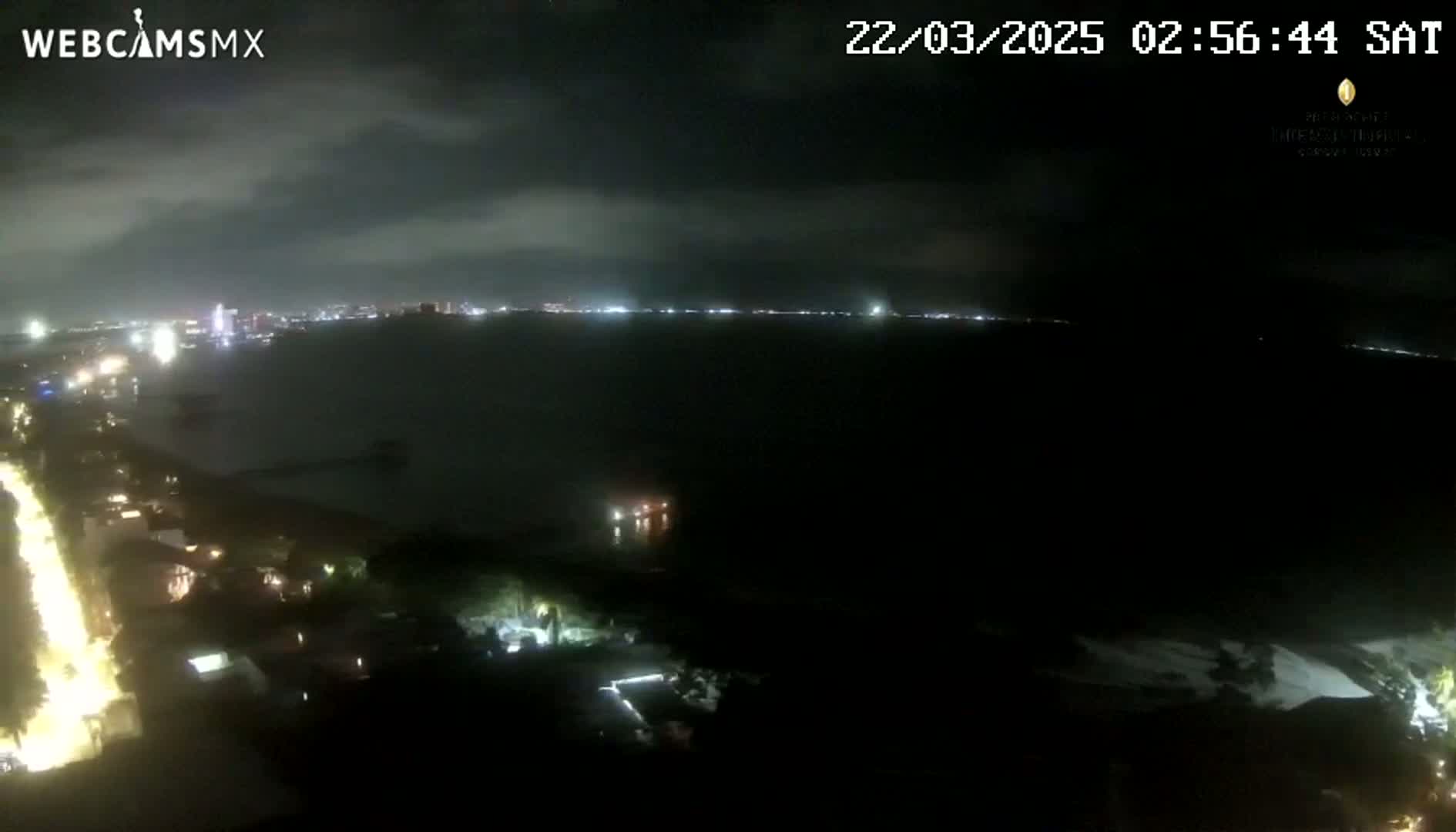 A nighttime, elevated view shows a dark body of water reflecting city lights on the horizon under a cloudy sky.