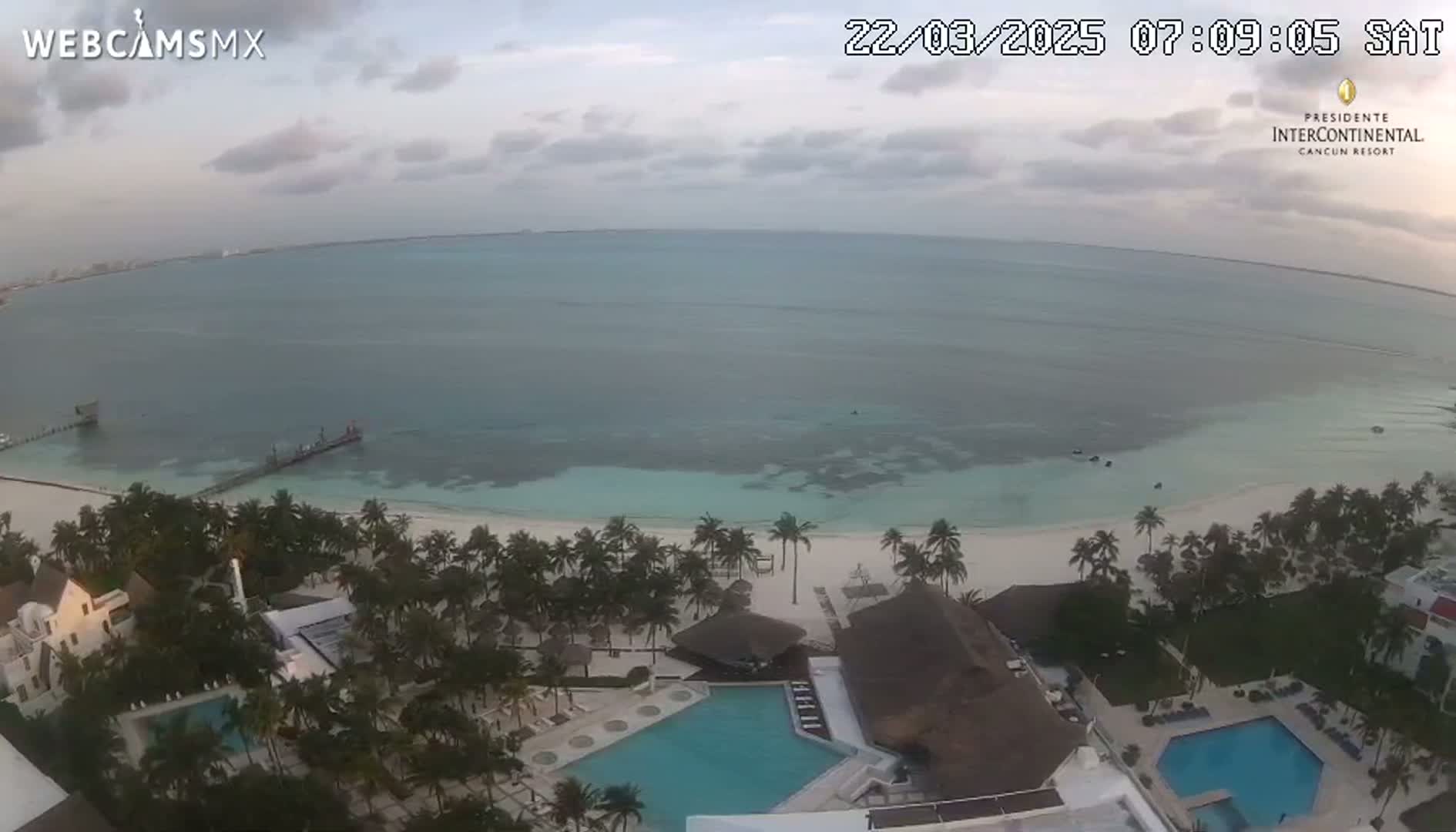 An aerial view shows a beach resort with pools and palm trees next to a calm, light-blue ocean under a mostly cloudy sky.