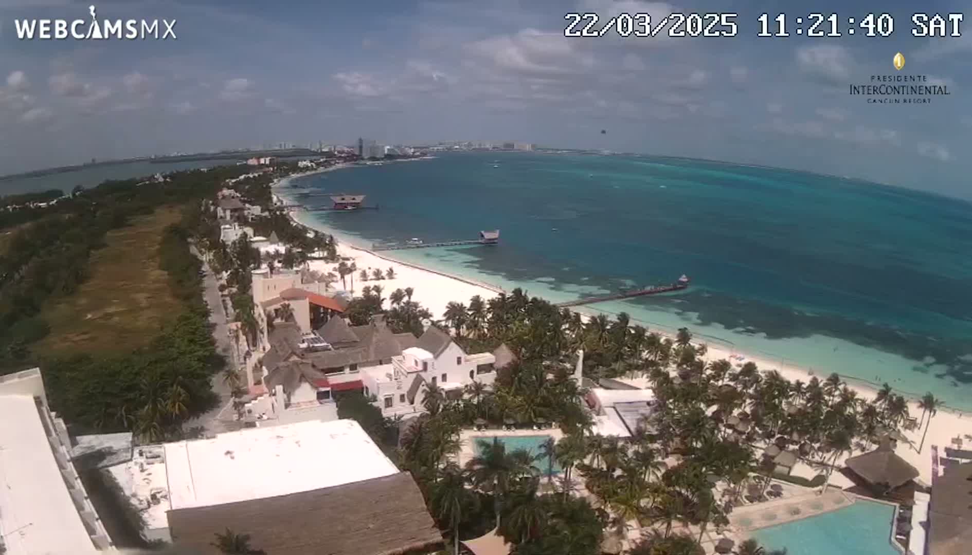 An aerial view shows a beach with turquoise water under a partly cloudy sky, featuring a resort with palm trees and swimming pools near the shore.