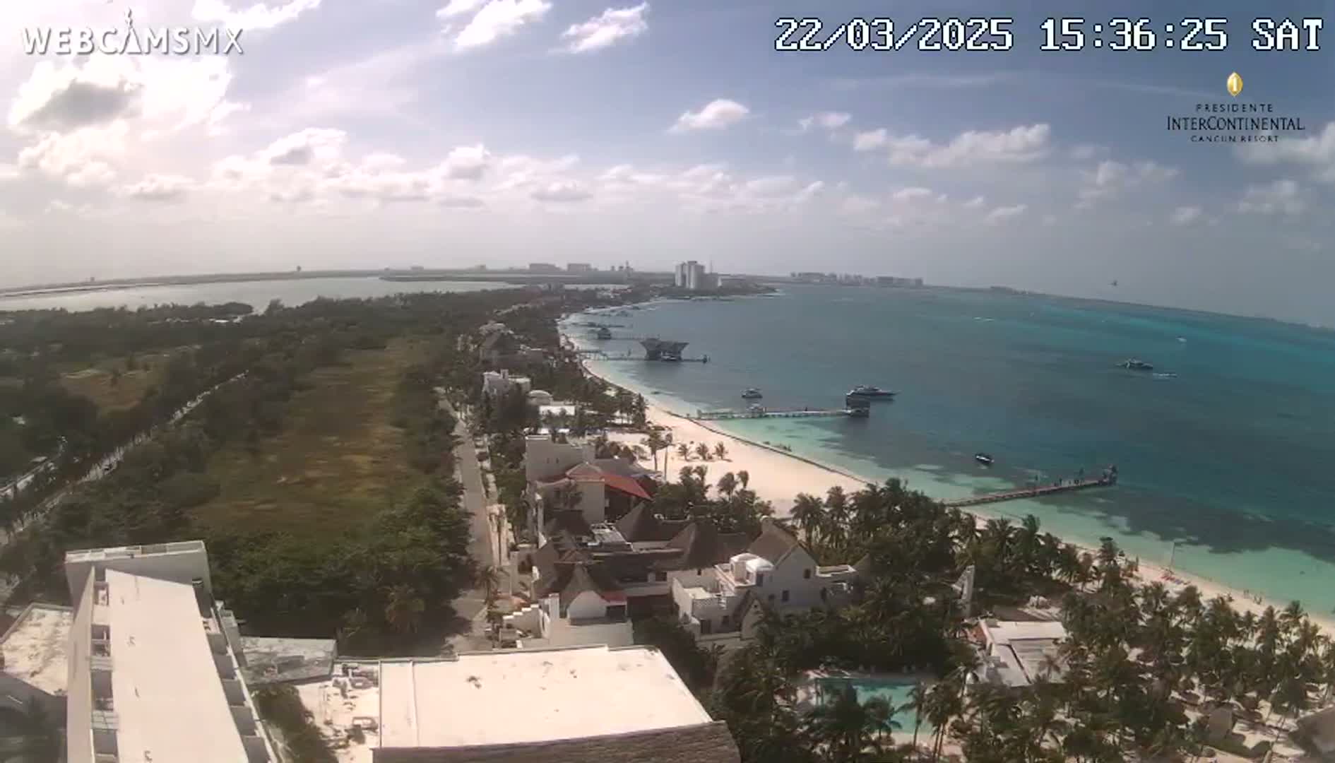 An aerial view shows a partly cloudy day over a tropical beach with white sand, turquoise water, palm trees, and beachfront buildings.