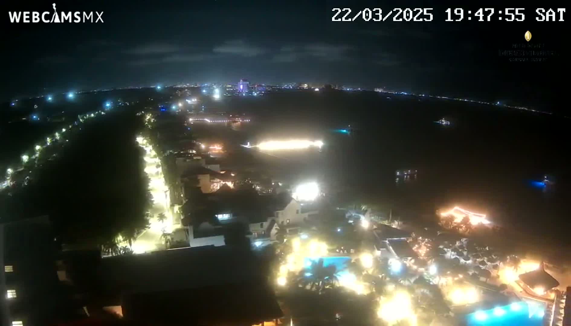 A nighttime, high-angle view shows a coastal resort town with brightly lit buildings and pools near a dark body of water under a mostly clear sky.
