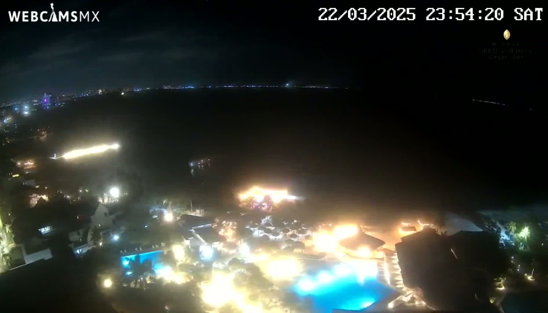 A nighttime aerial view shows a resort complex with illuminated pools and buildings near a dark body of water under a clear sky.
