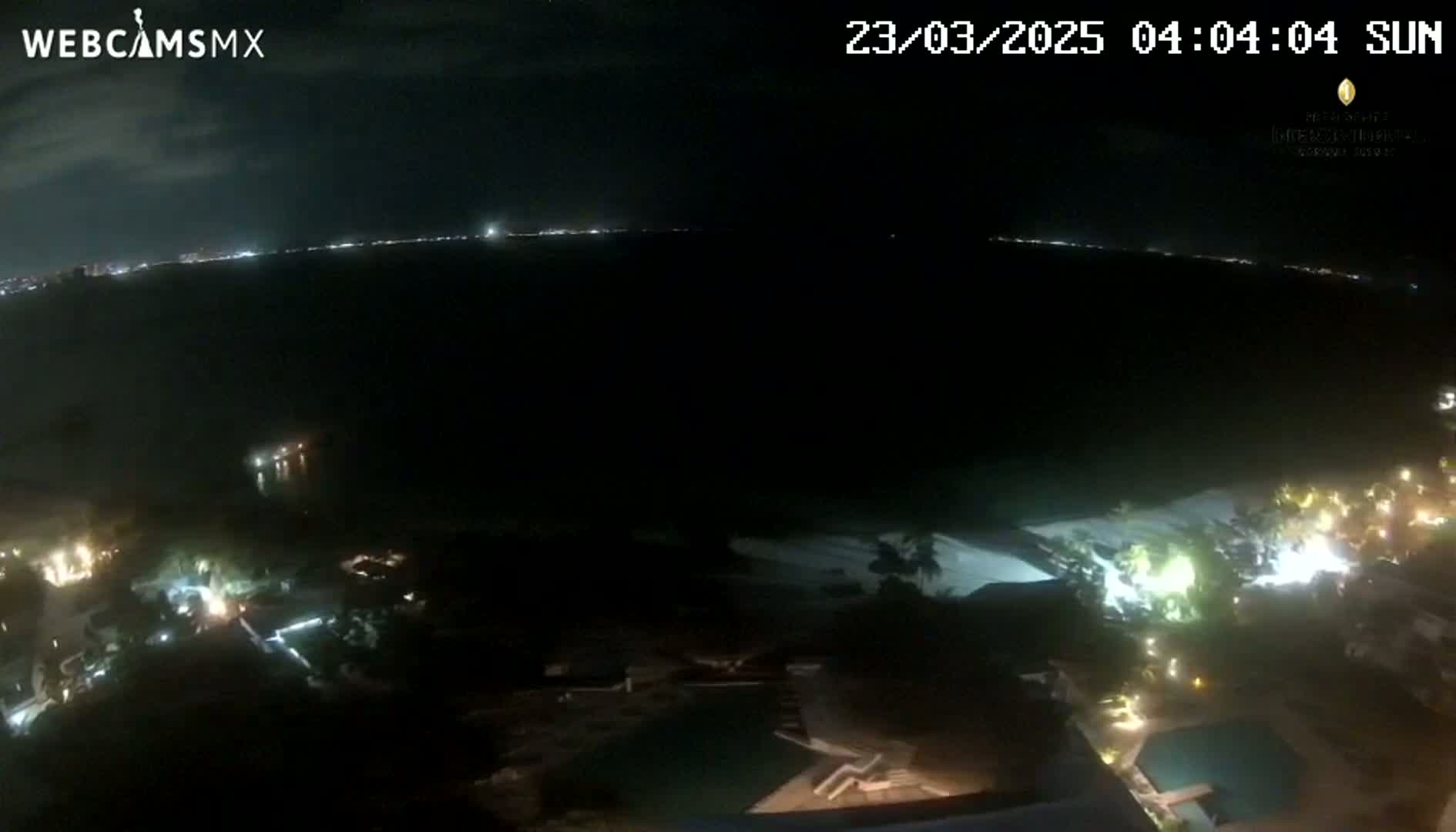A nighttime aerial view shows a dark body of water with a distant city skyline illuminated, and a resort area with pools below, under a clear, dark sky.
