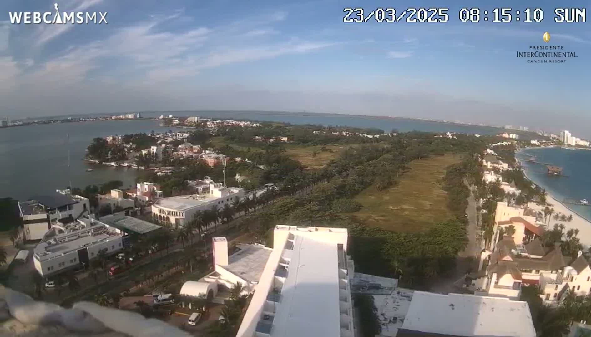 A high-angle, sunny view shows a coastal city with a large body of water, buildings, and vegetation, under a mostly clear sky.