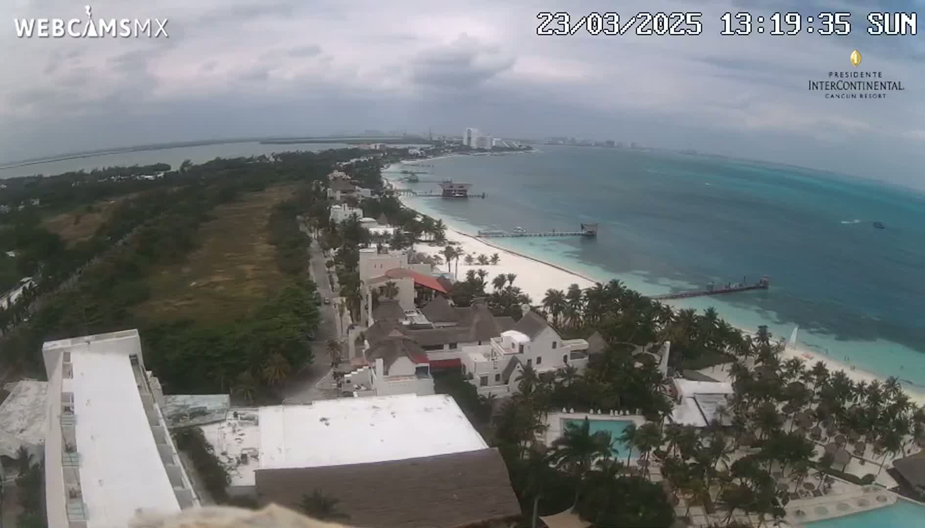 An aerial view shows a tropical beach with resorts, palm trees, and overcast skies.