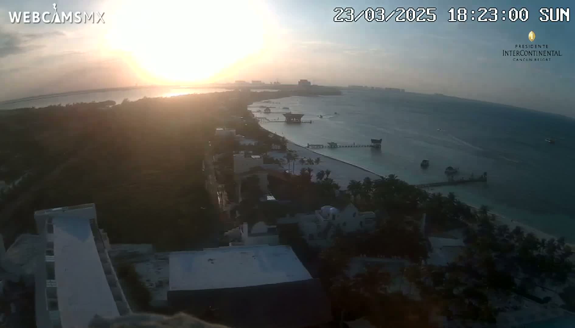 A high-angle view shows a sunset over a calm body of water next to a beachfront area with buildings and palm trees, during what appears to be a clear evening.