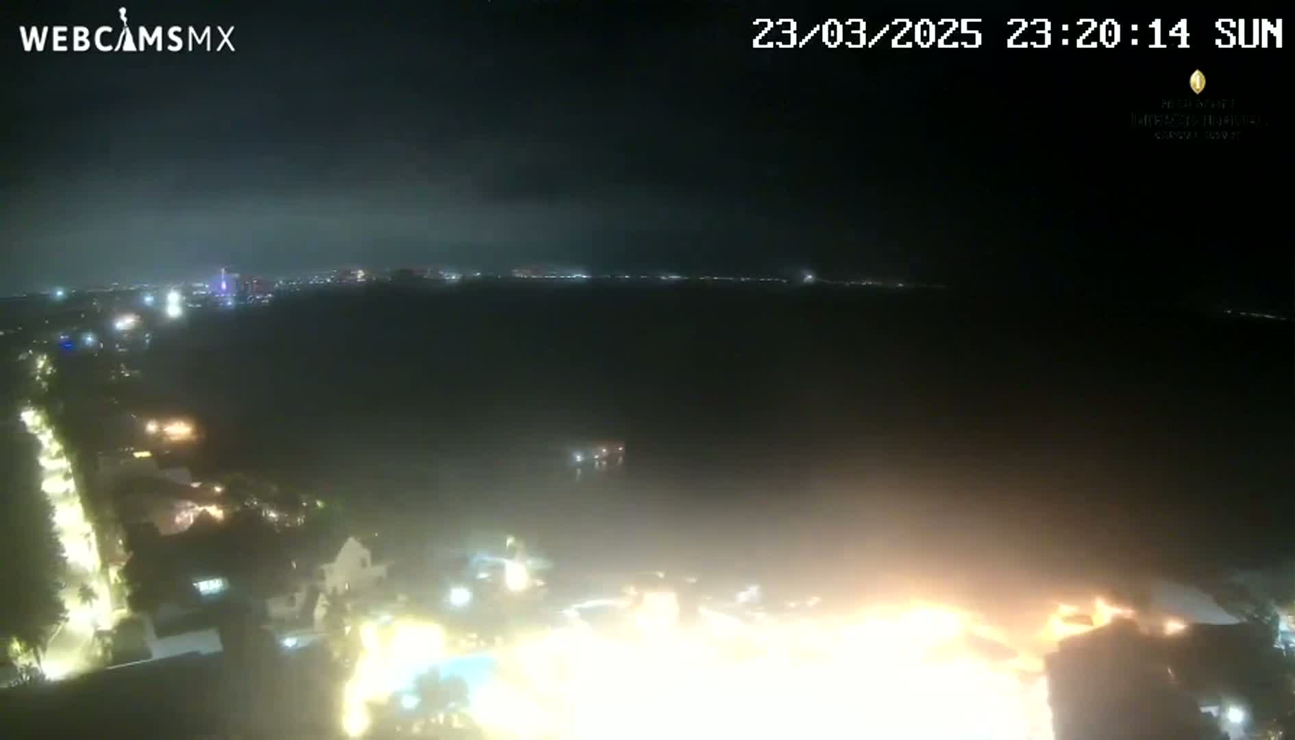 A nighttime, aerial view shows a body of water with city lights on the horizon, illuminated buildings and streets below, under a mostly clear sky.