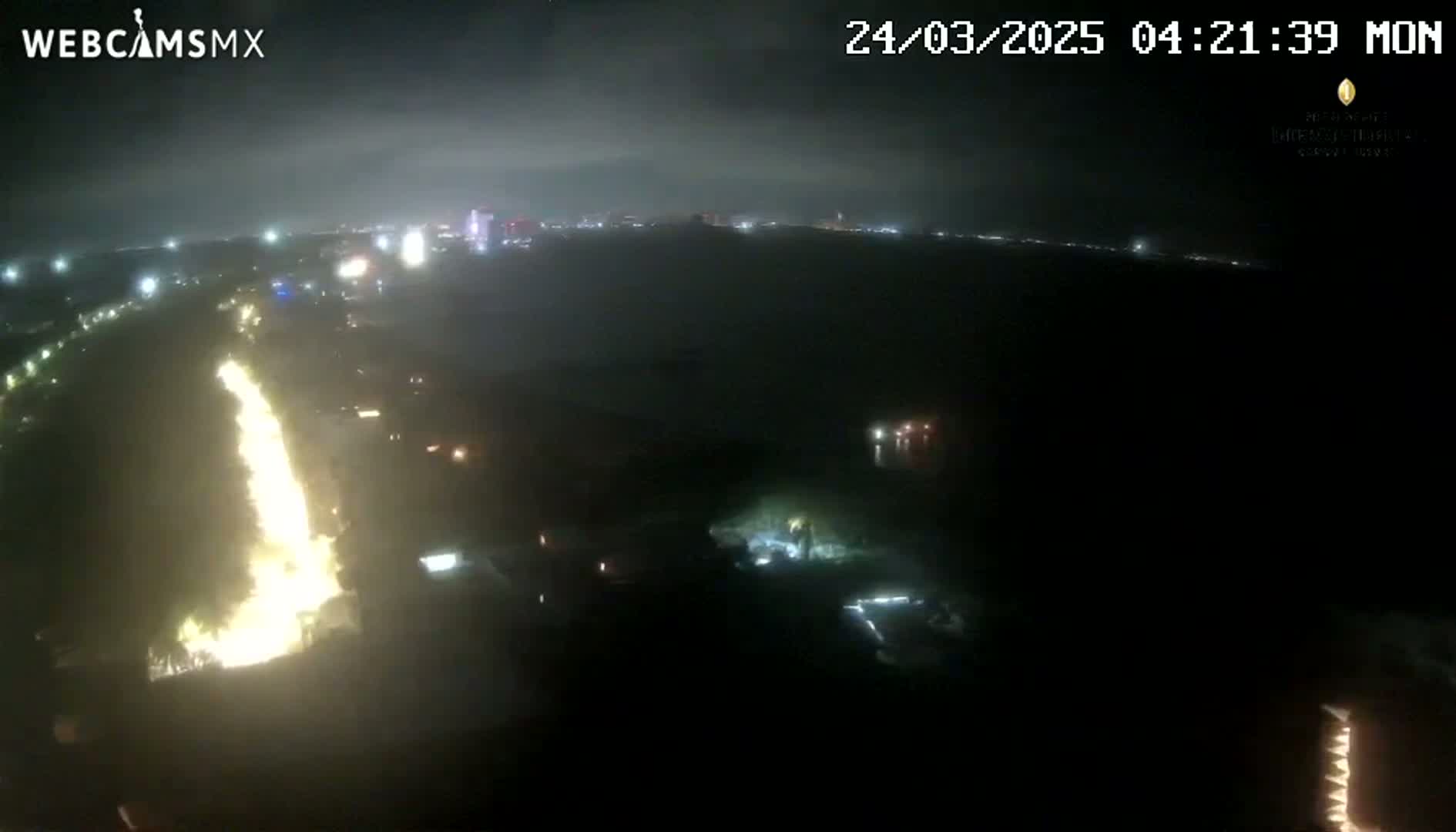 A nighttime aerial view shows a bright, vertical streak of light, possibly a fire, near a dimly lit city under a cloudy sky.