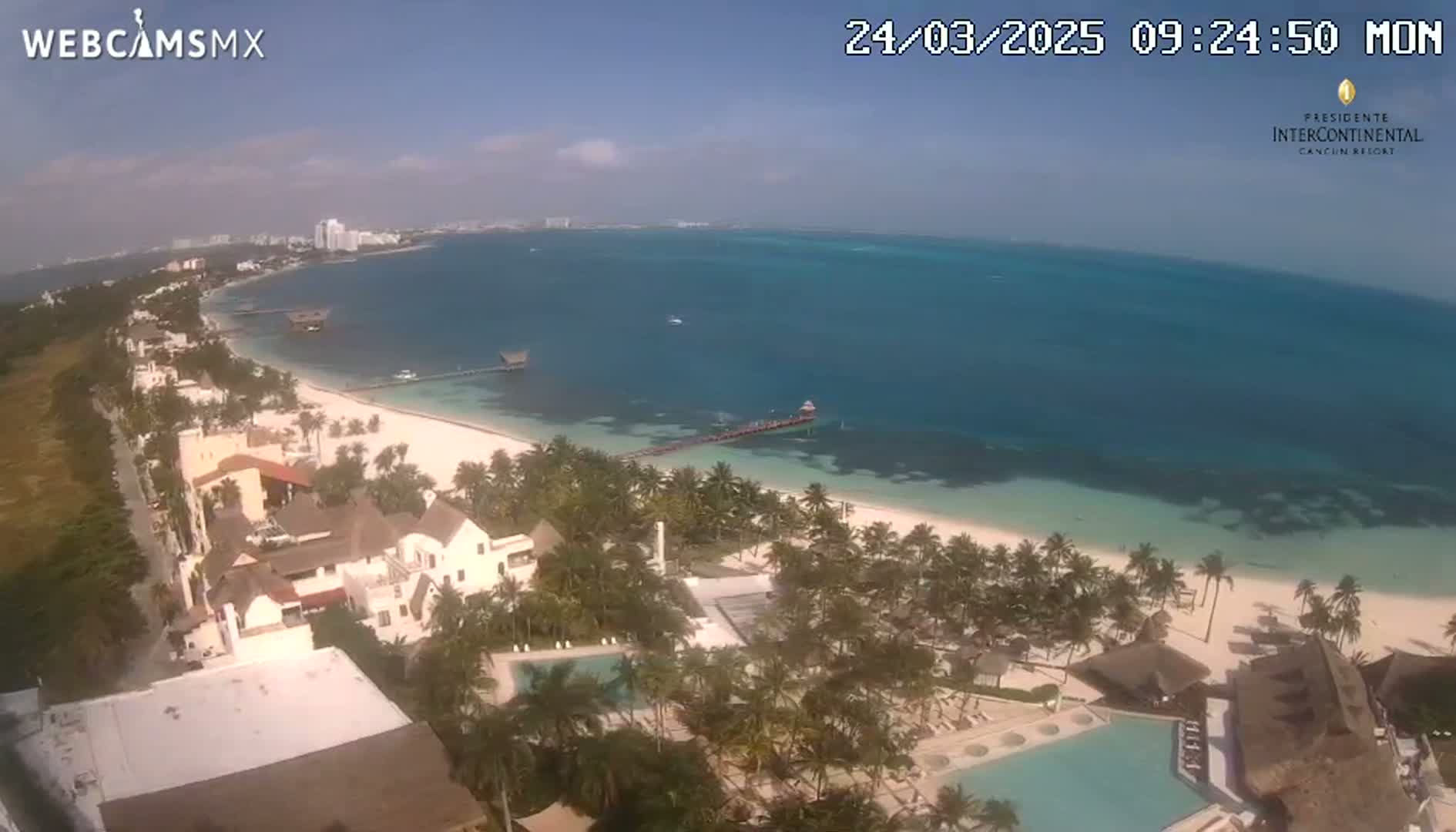 An aerial view shows a beach with turquoise water under a clear, sunny sky, with palm trees, buildings, and swimming pools lining the coast.