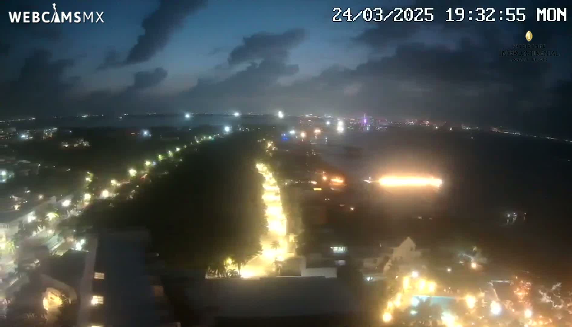 A nighttime, aerial view of a coastal city with a brightly lit road winding through the darkness, under a partly cloudy sky.