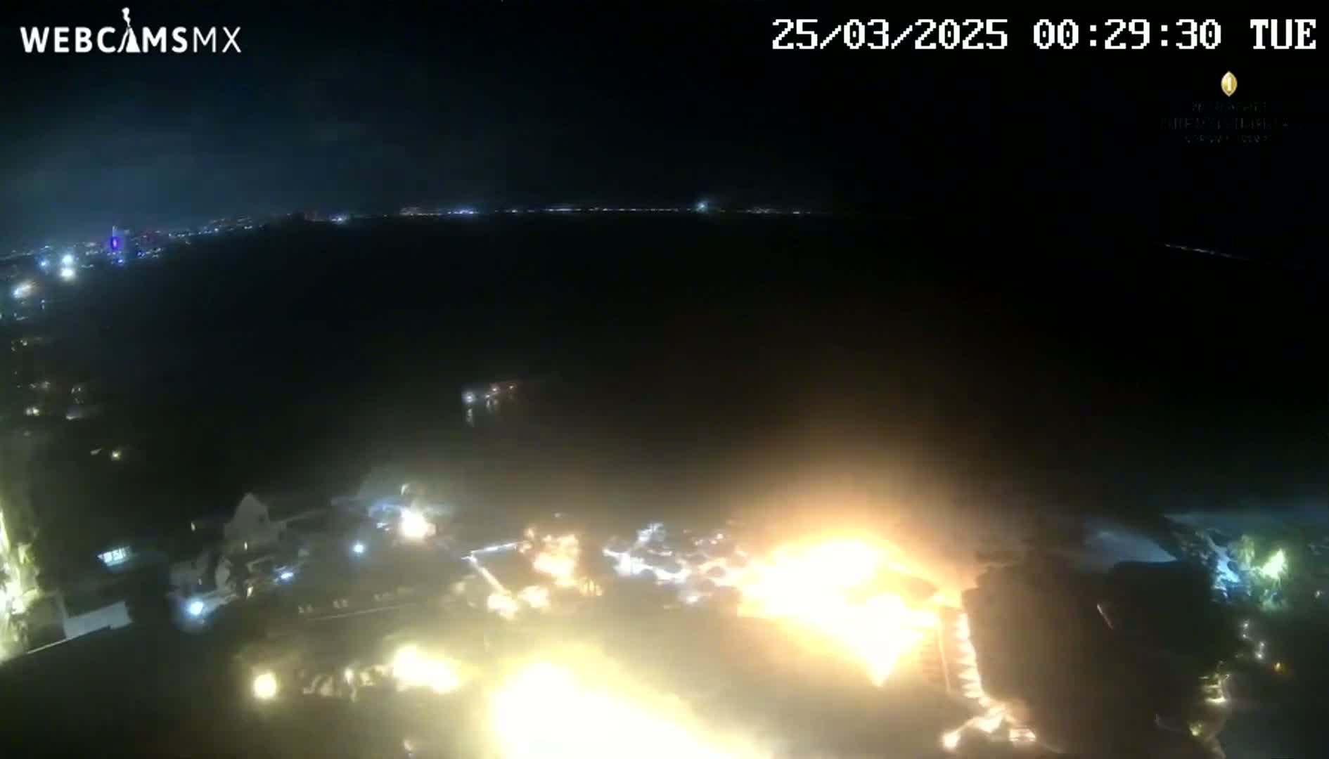 A nighttime aerial view shows a large body of water with bright lights and a significant fire or explosion near the shore, under a dark, clear sky.