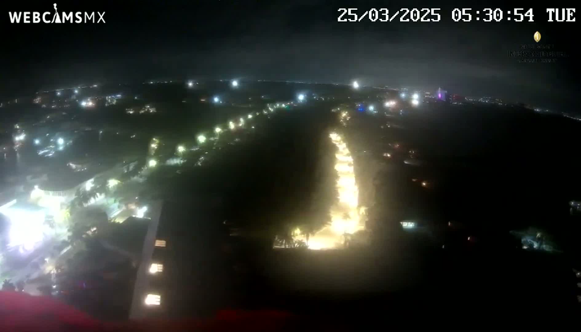An aerial nighttime view of a city shows a brightly lit road winding through a darker, tree-filled area under a cloudy sky.