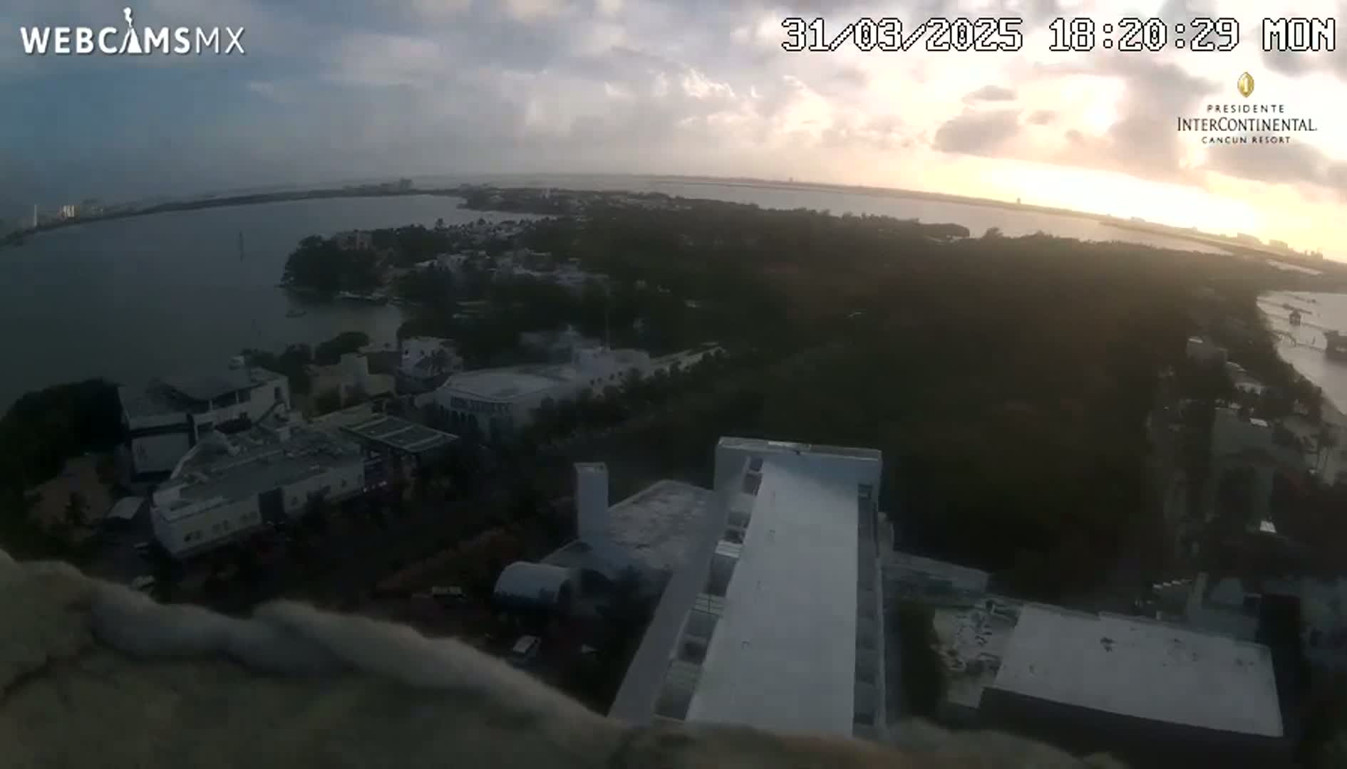 An elevated view shows a body of water at sunset, with buildings and land visible below, under partly cloudy skies.