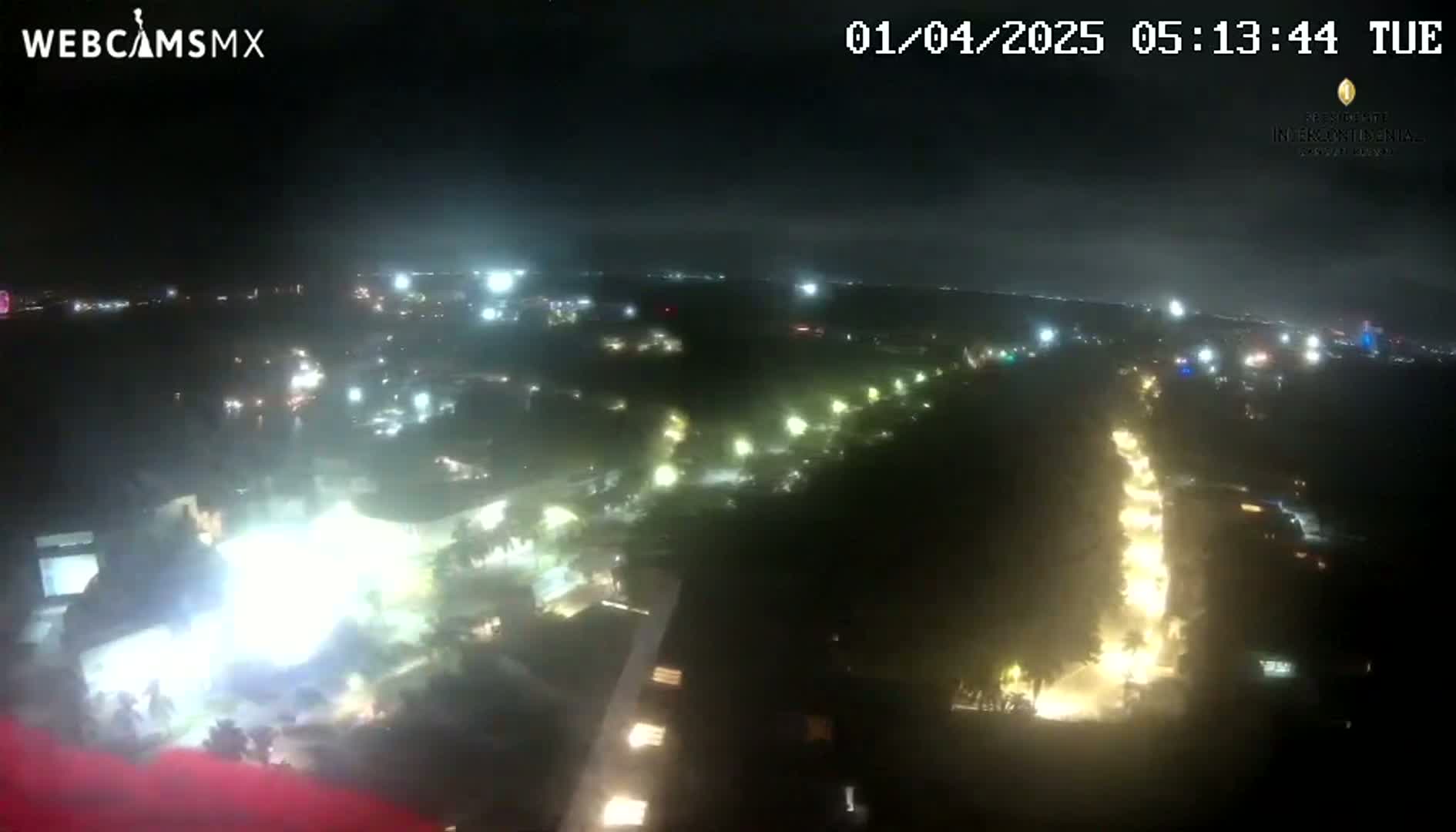 A nighttime aerial view shows a sprawling area with numerous lights illuminating buildings and roads under a mostly cloudy sky.